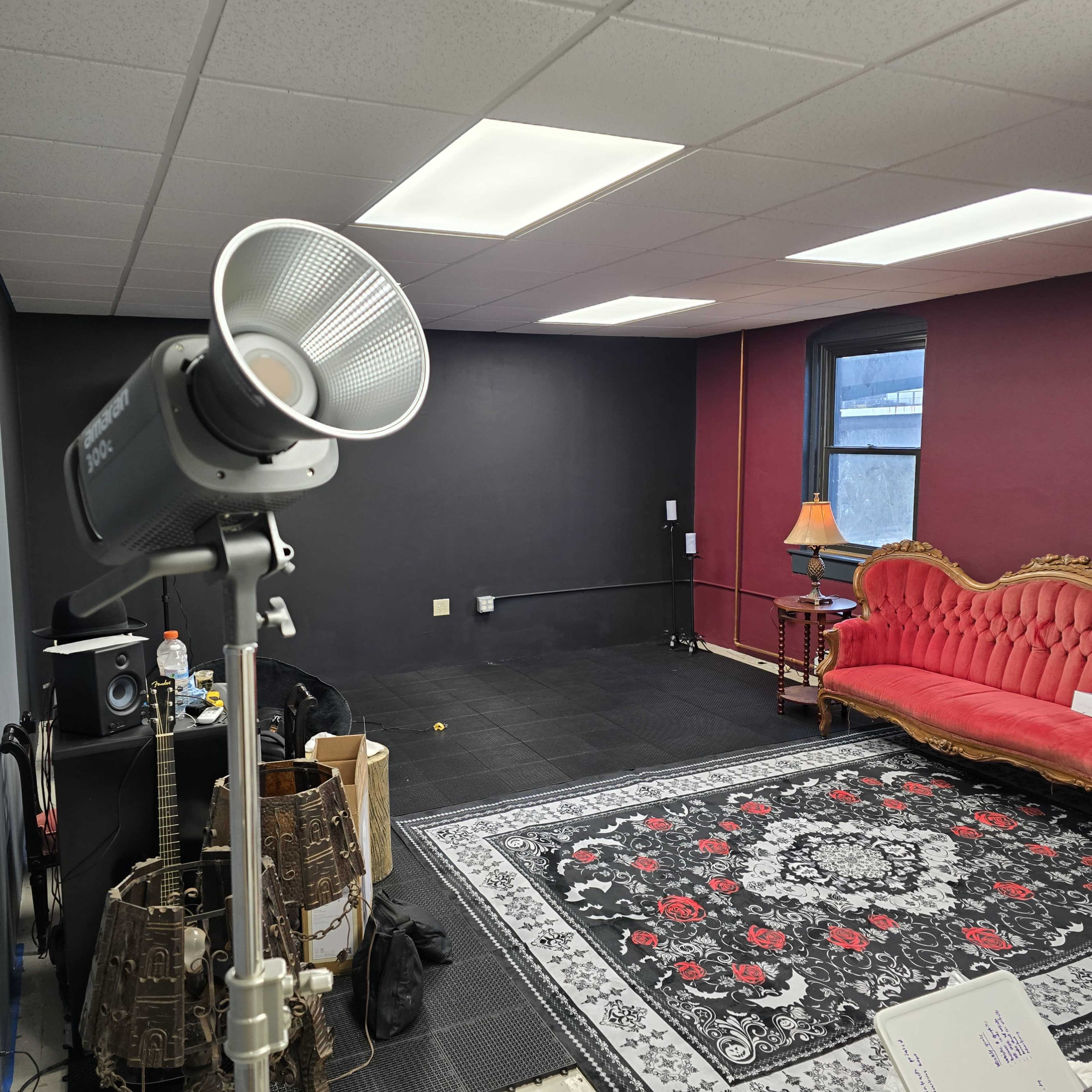 The image shows a room with a red velvet couch, a lamp, a patterned rug, and a lighting setup in one corner.