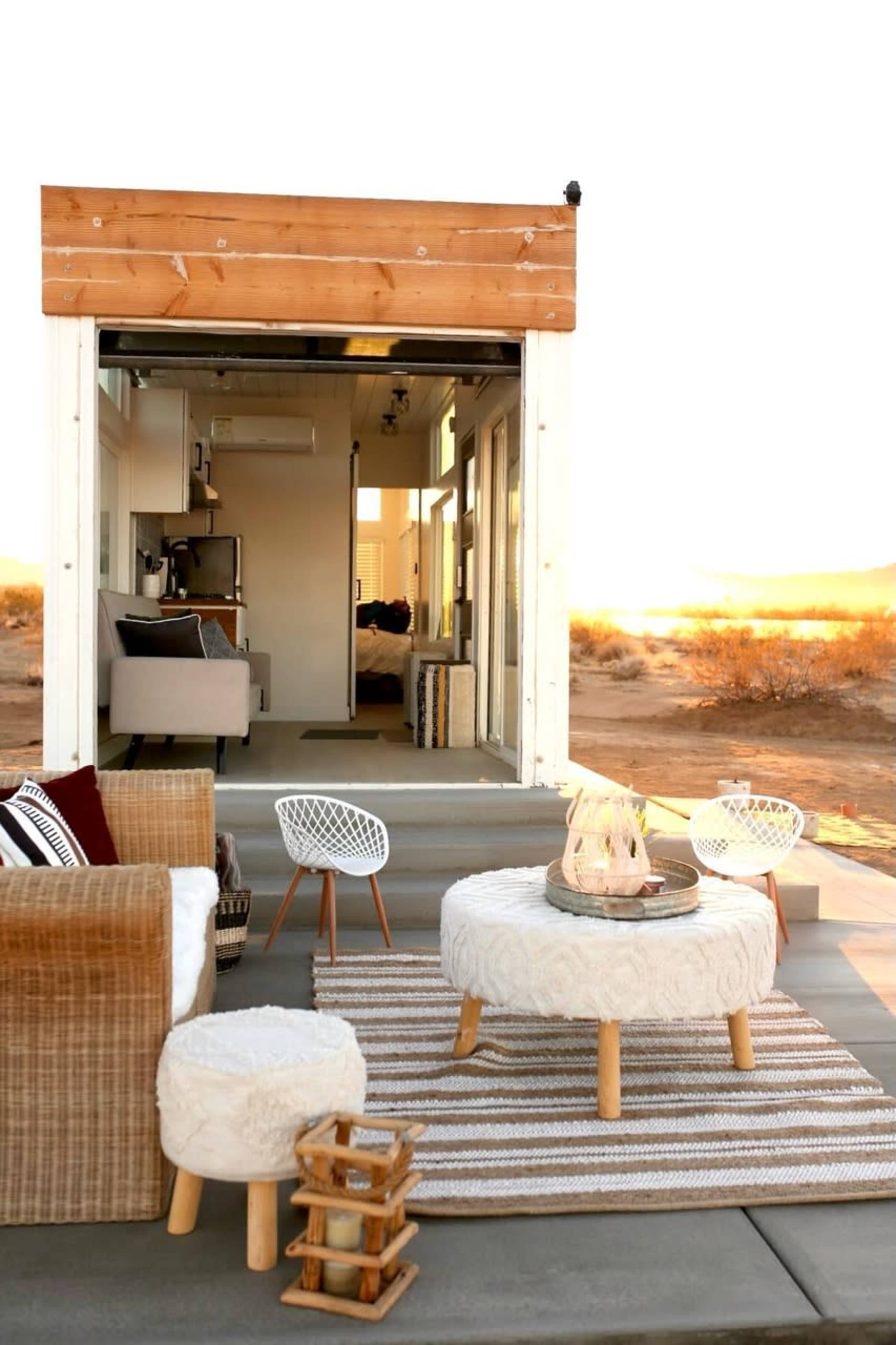 A cozy outdoor seating area features a round coffee table, woven chairs, and a striped rug in front of a tiny house with large sliding doors.