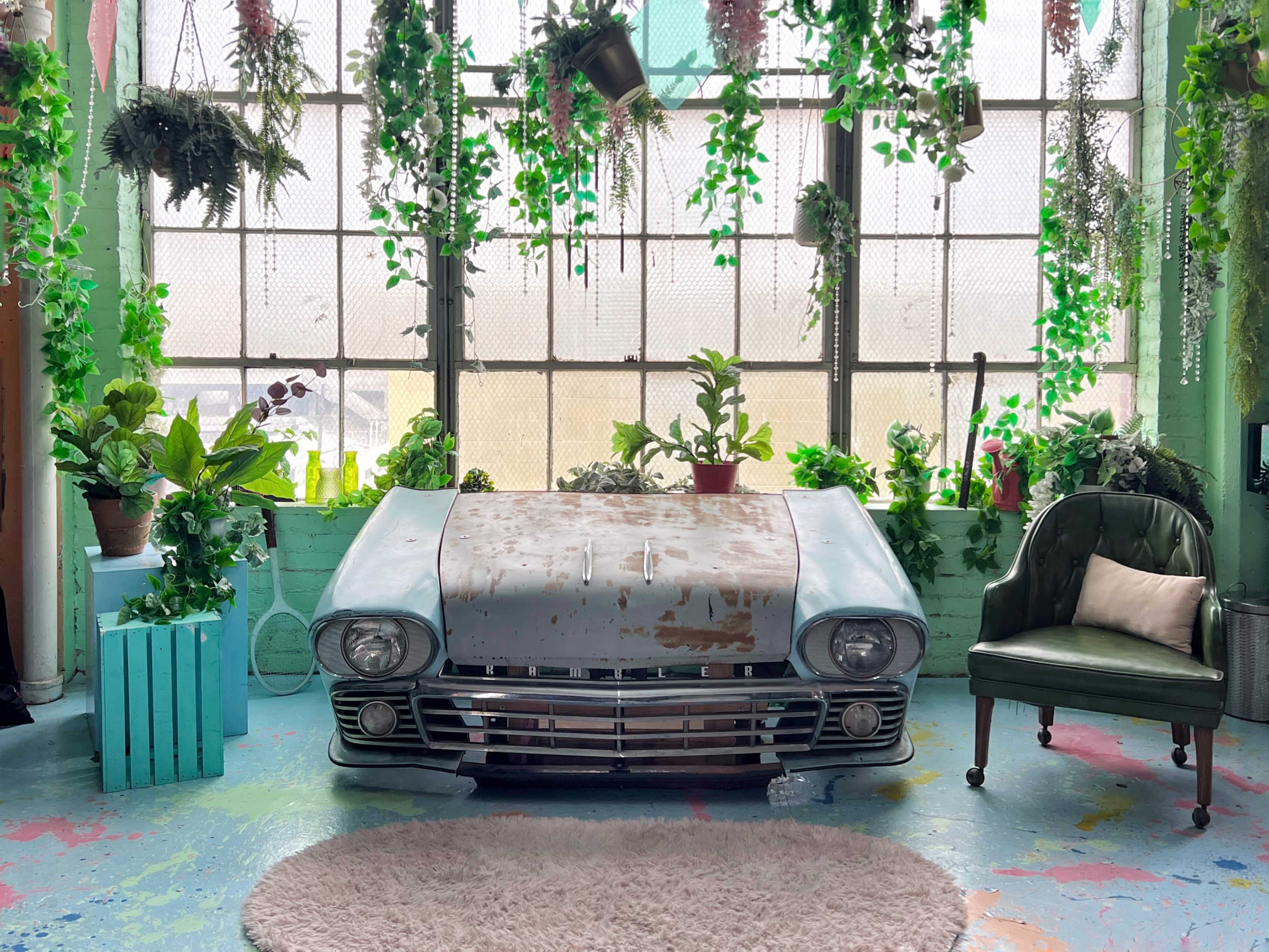 The image features an old, rusted car hood placed on the floor of a room filled with various indoor plants and greenery in front of a large window.