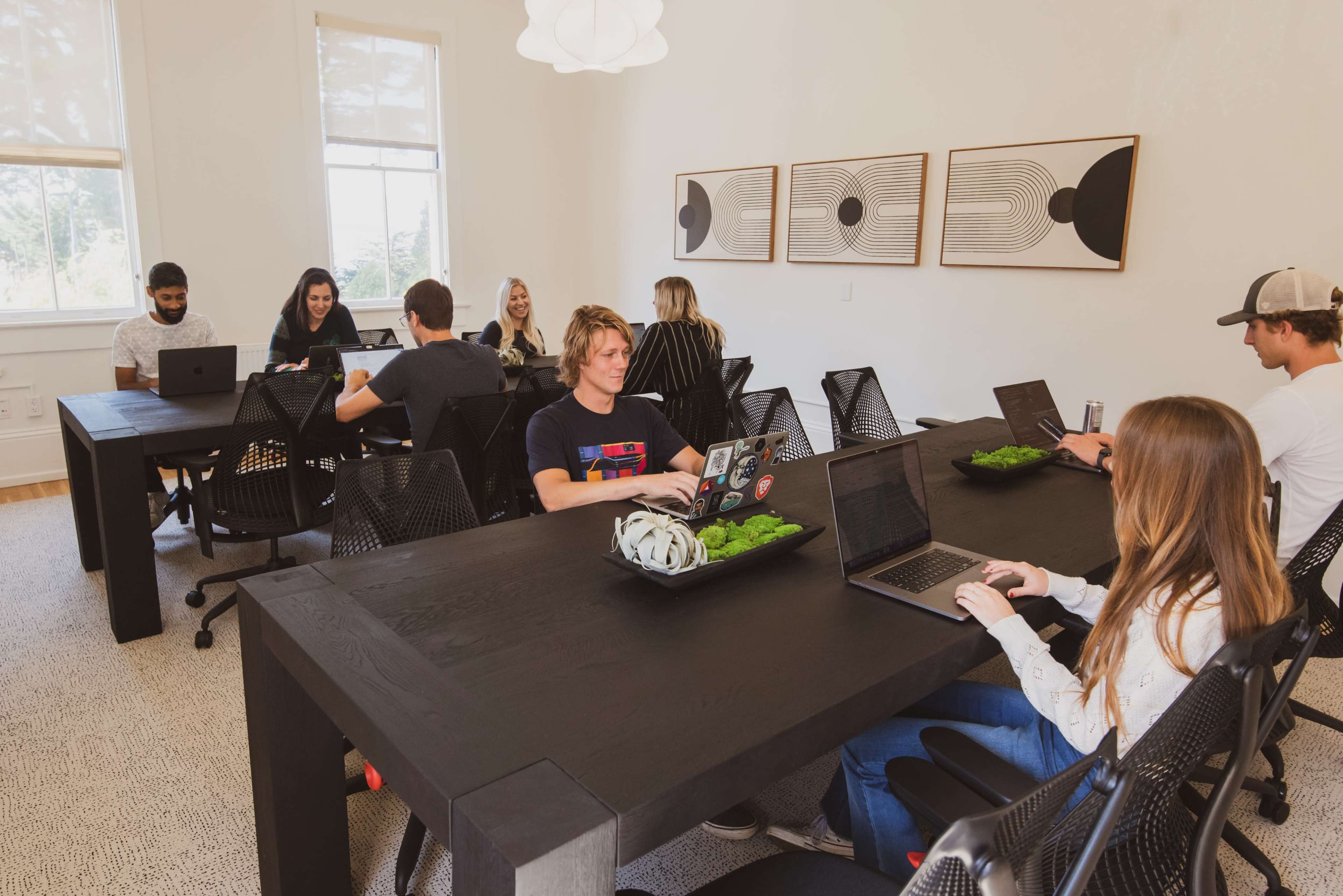 A group of people works on laptops at a large conference table in a bright office space.