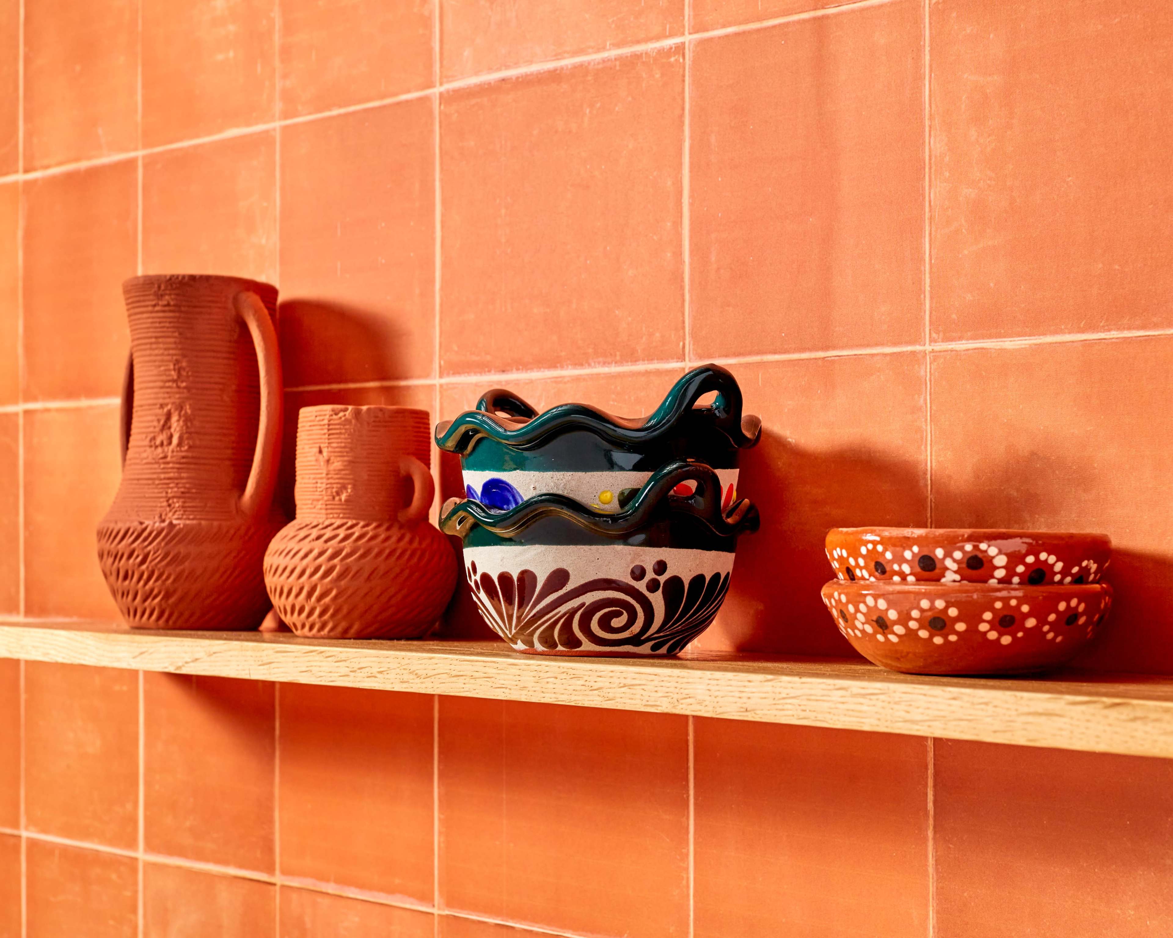 A wooden shelf displaying a variety of colorful ceramic bowls and clay pitchers against an orange tiled backdrop.