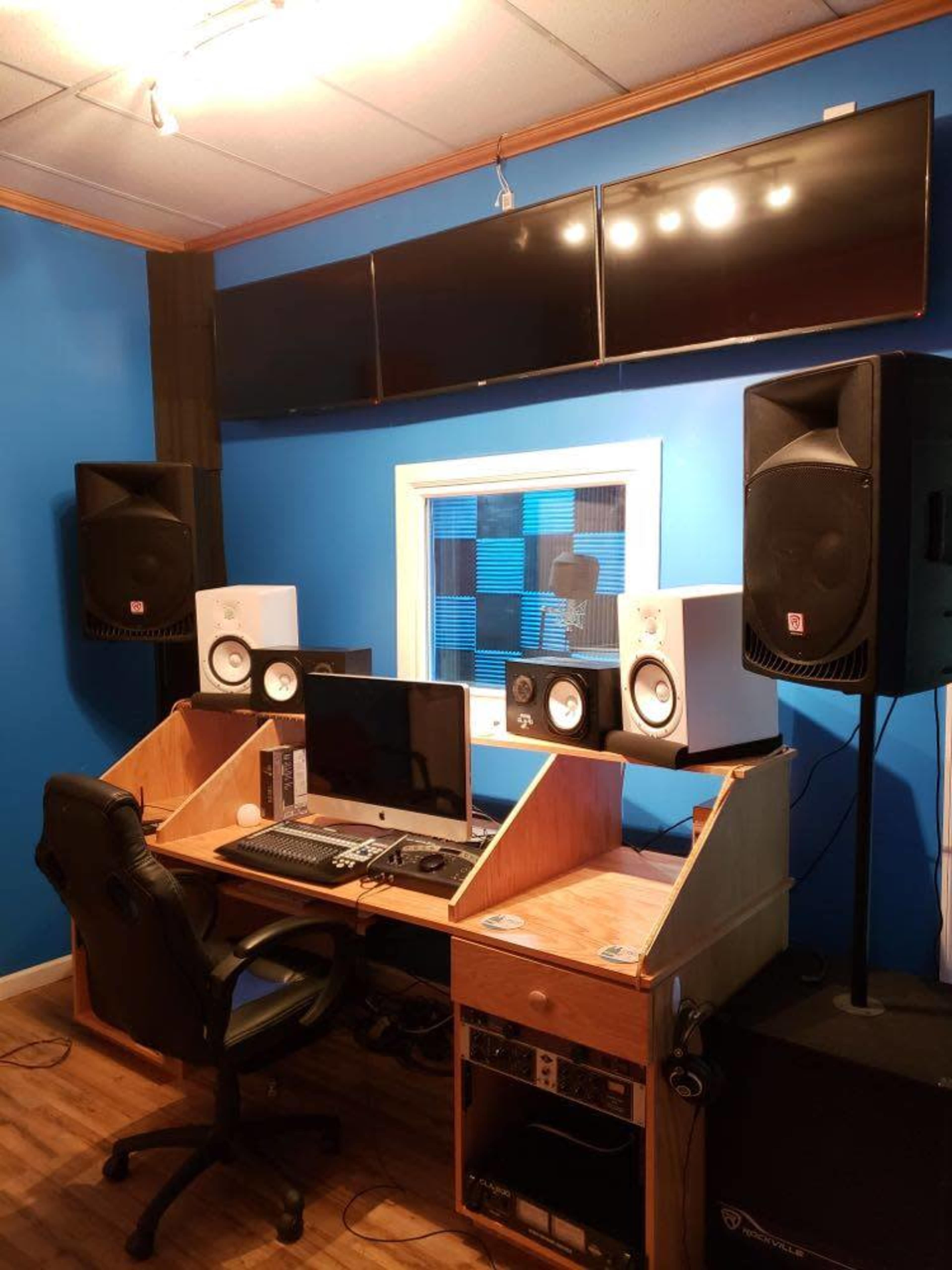 The image shows a music studio with a computer desk, audio monitors, and multiple screens mounted on a blue wall.