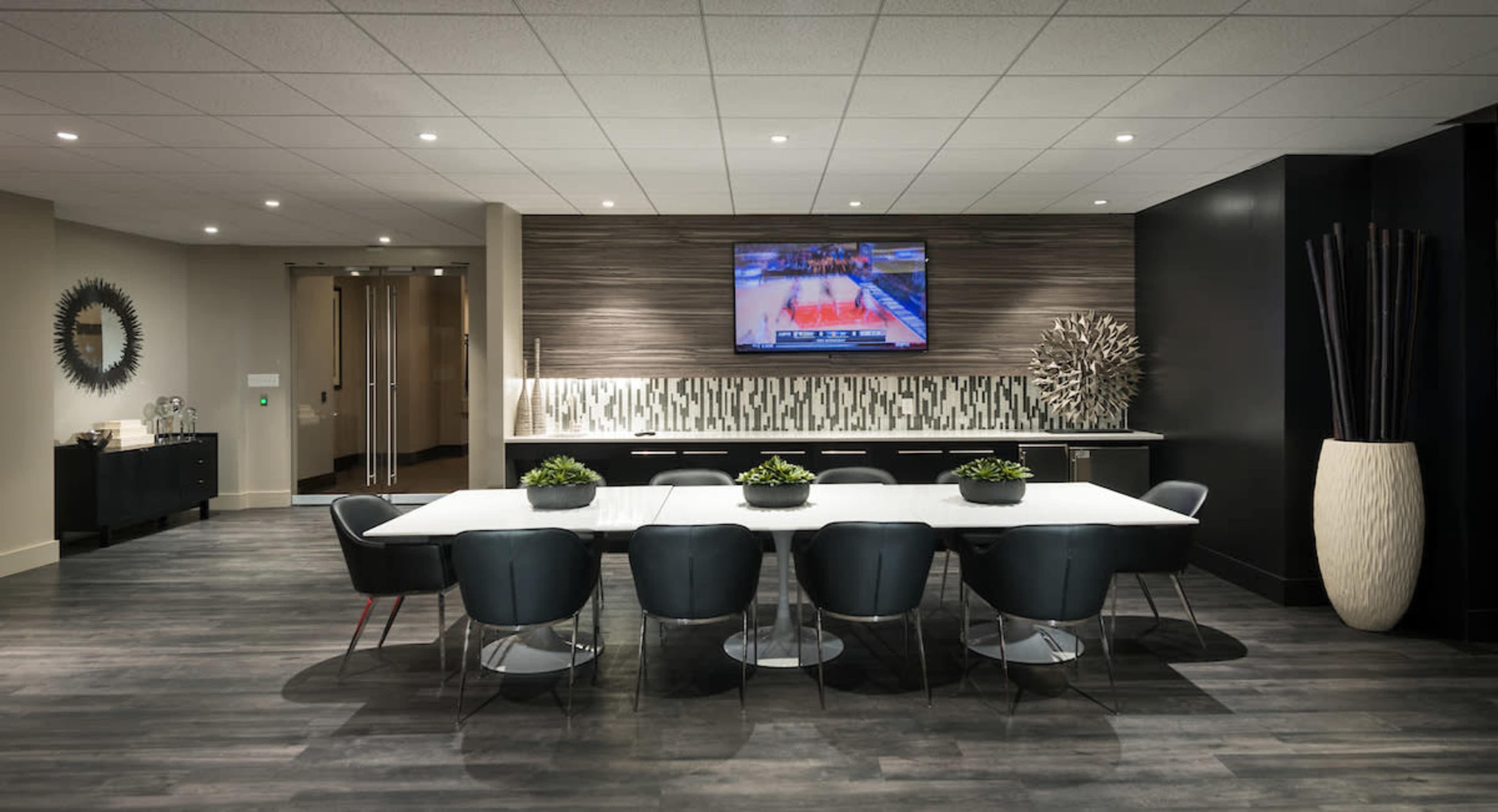 A modern conference room features a long white table surrounded by black chairs, with a television displaying a basketball game on the wall.