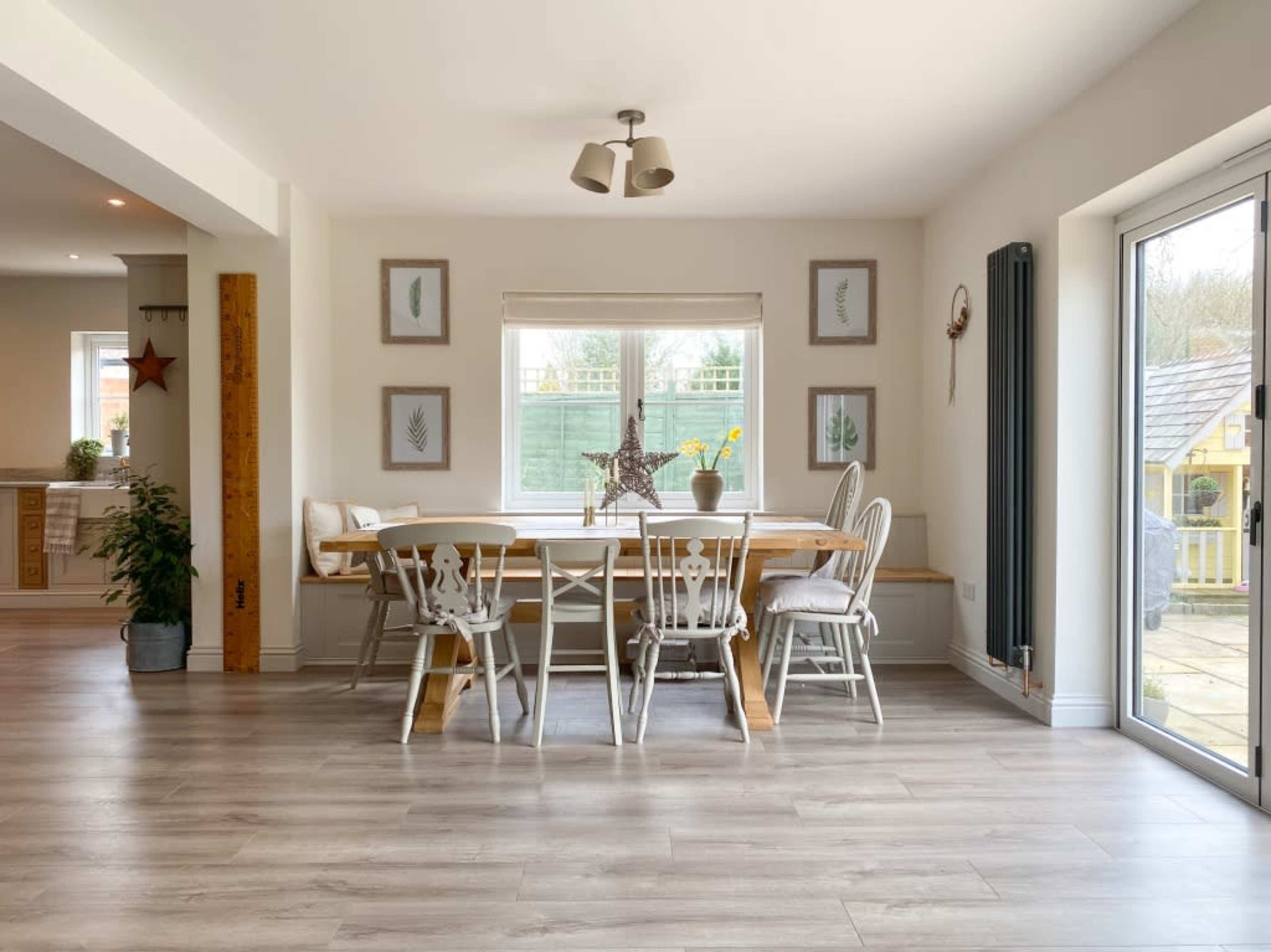 A bright dining area features a wooden table surrounded by chairs, with a window showing greenery outside and framed plant artwork on the walls.