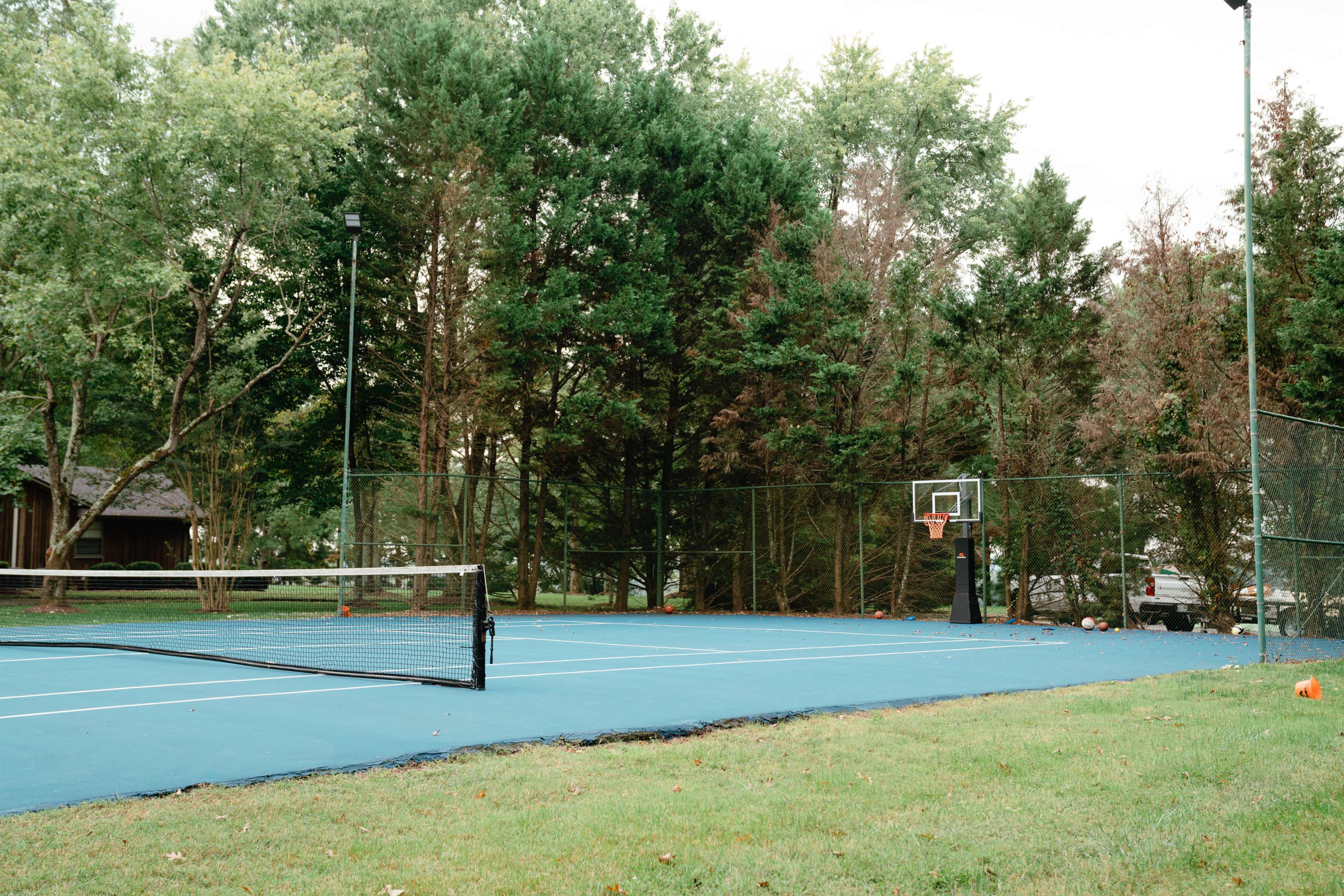 A blue court is set up for tennis and basketball, surrounded by trees and grass.