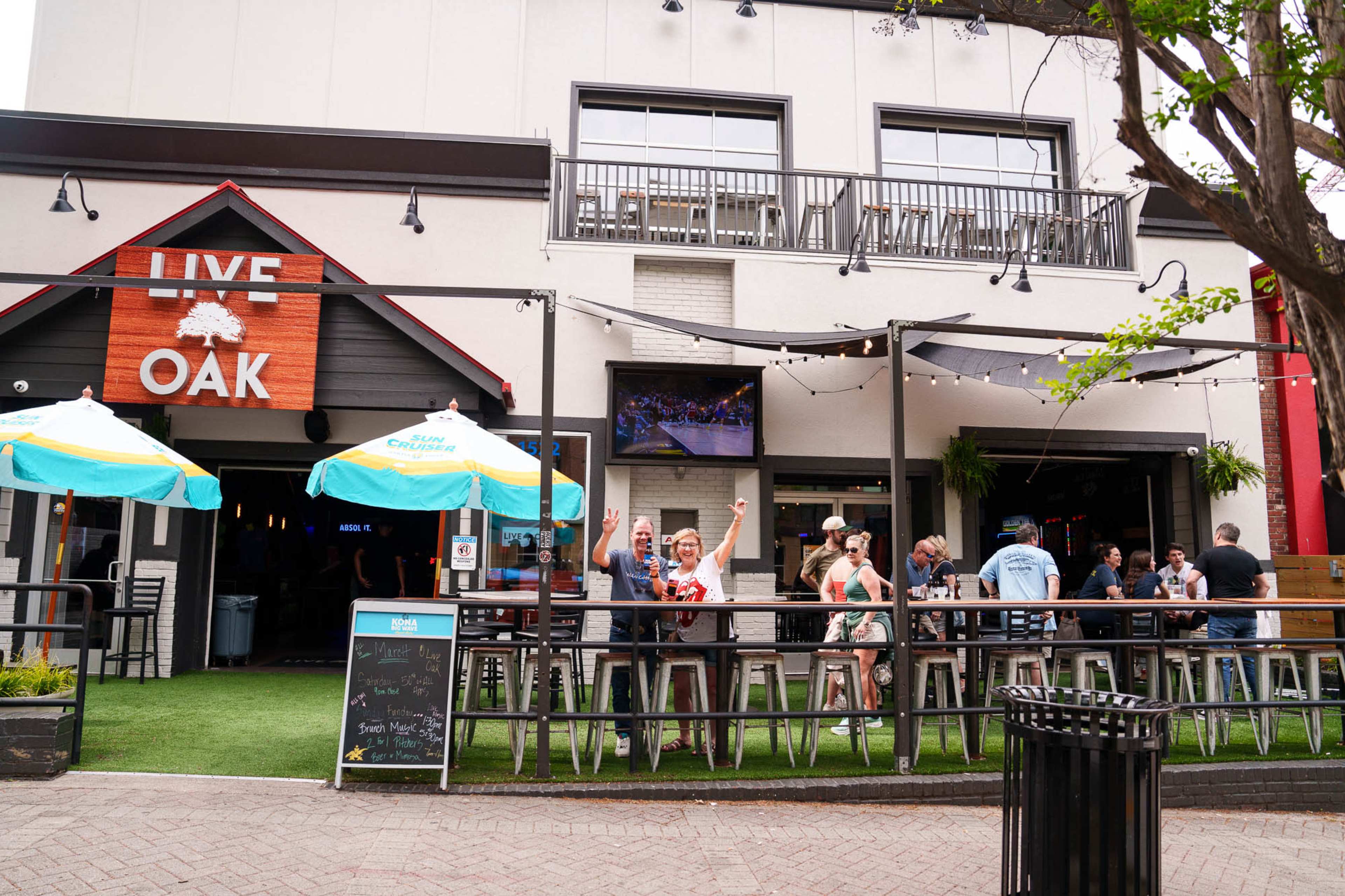 The image shows a lively outdoor bar area with patrons enjoying drinks under umbrellas outside a building marked "LIVE OAK."