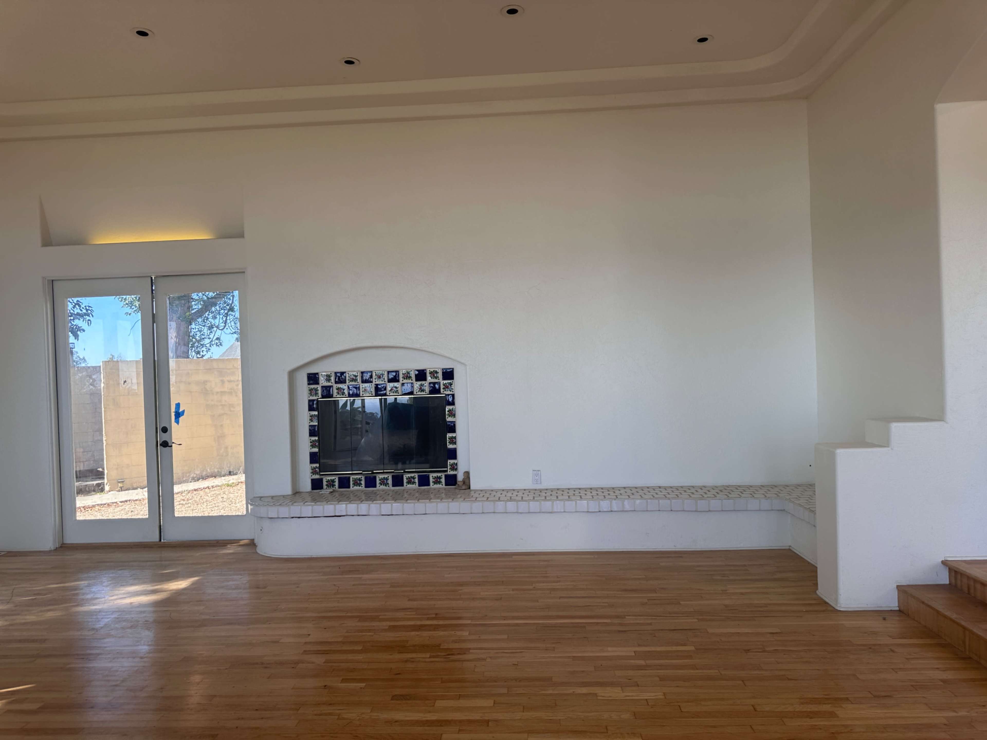 The image shows a spacious, empty room with wooden flooring, a fireplace featuring blue tiled accents, and large glass doors leading outside.