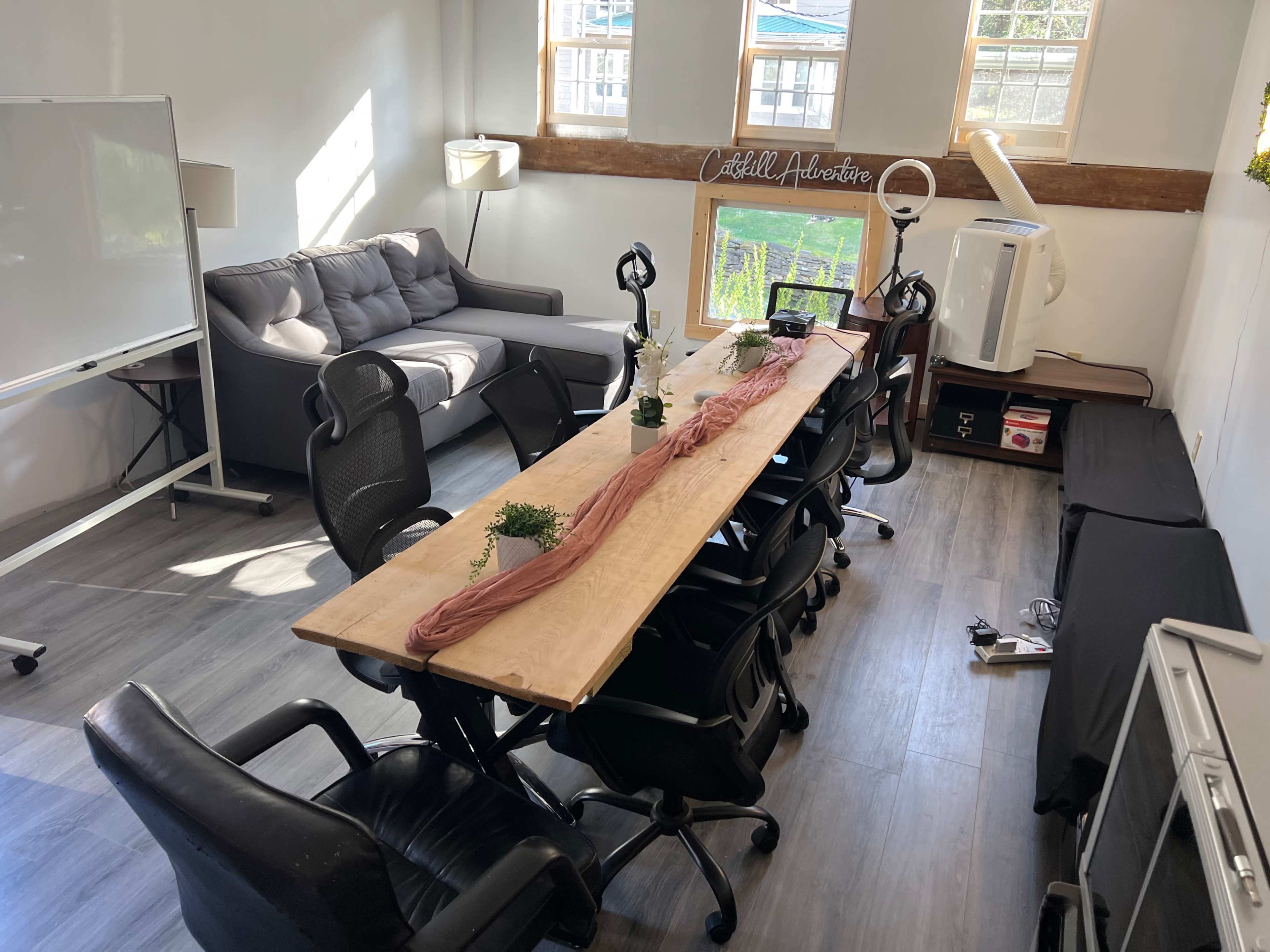 A modern meeting room features a long wooden table surrounded by black chairs, with a gray sofa and windows providing natural light.