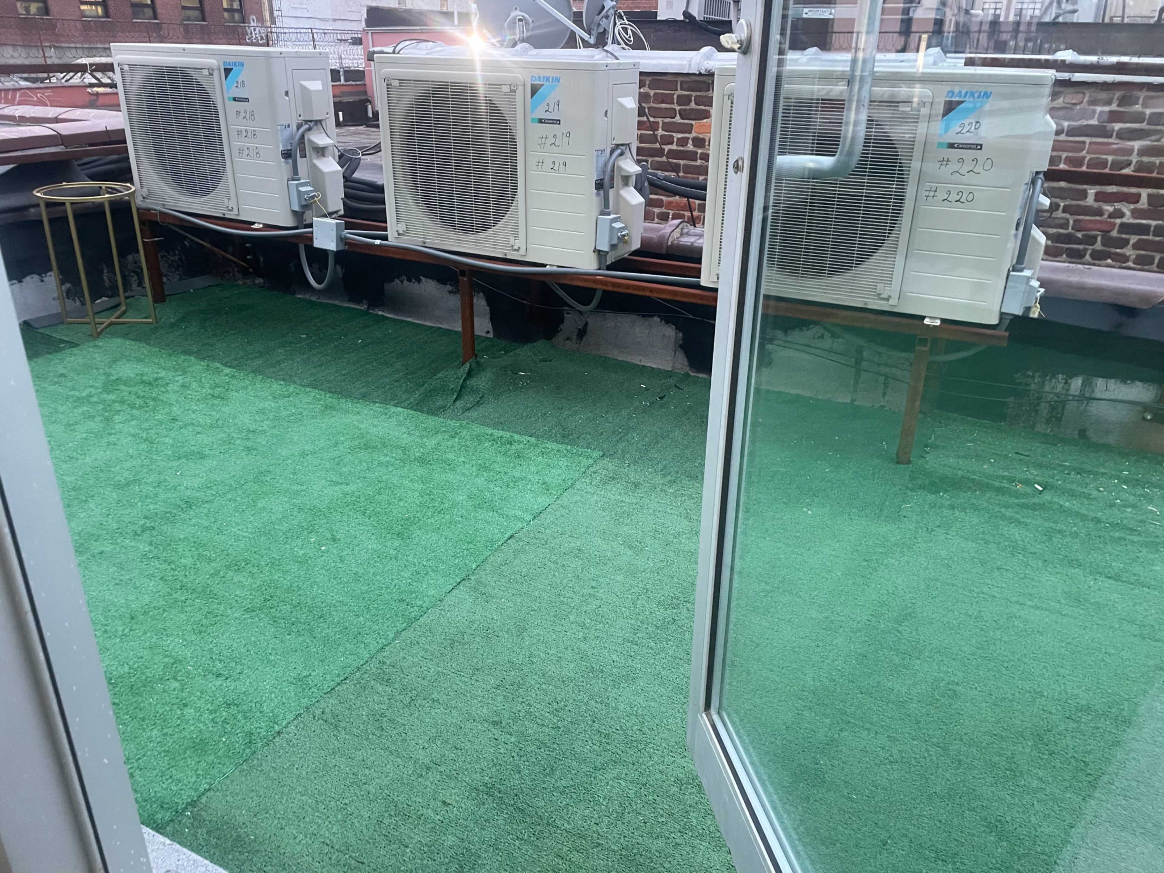 The image shows a rooftop area with three air conditioning units placed on a green artificial grass surface, along with a small gold-colored table near the edge.