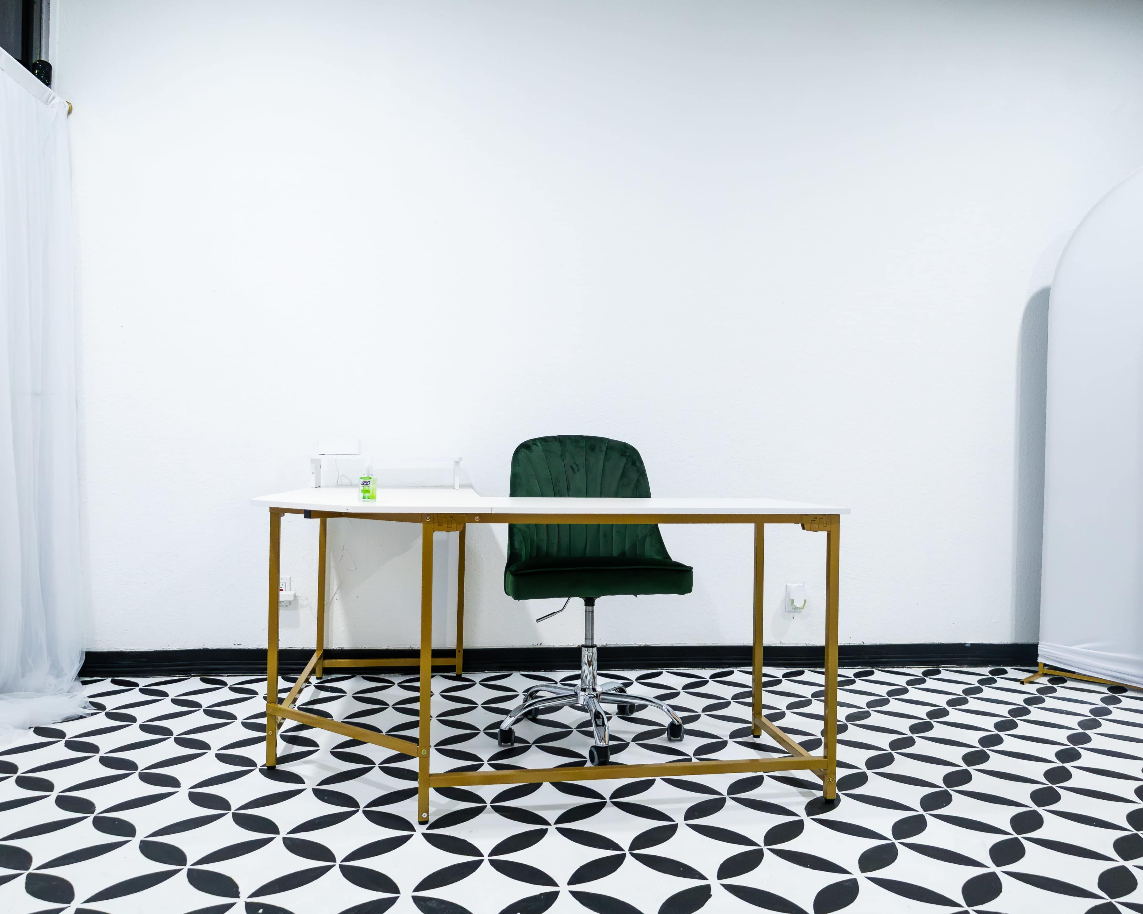 A green office chair is positioned at a white desk on a patterned black and white floor in a sparse room.