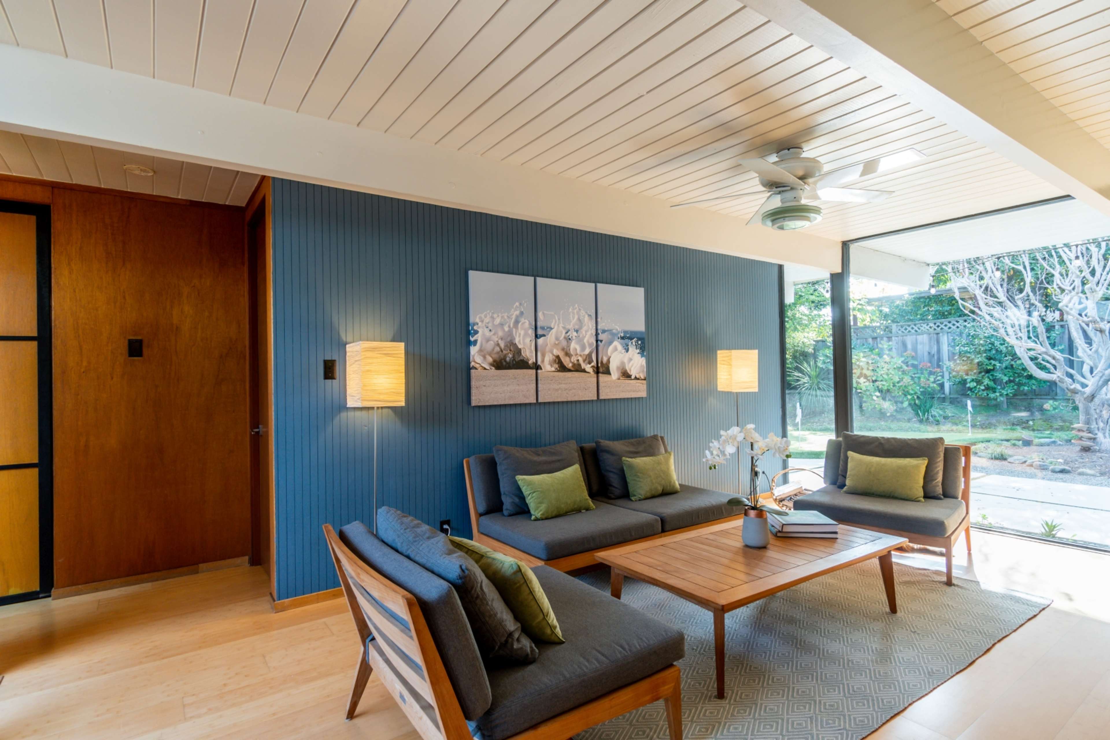 A modern living room with two gray sofas and a wooden coffee table, featuring framed artwork on the wall and a view of a garden through large glass doors.
