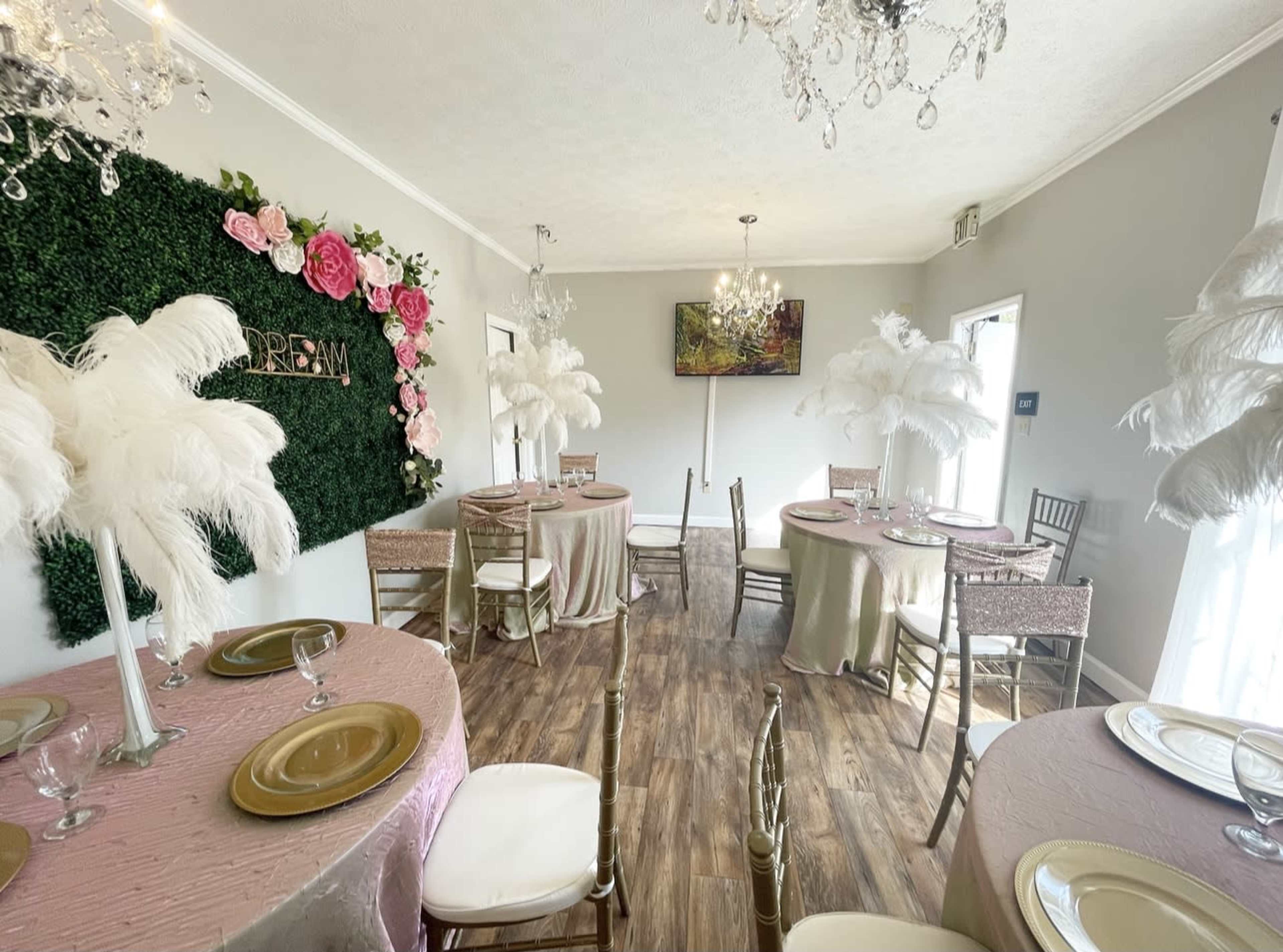 A decorated event space features round tables with elegant table settings, adorned with feather centerpieces and a floral accent wall.