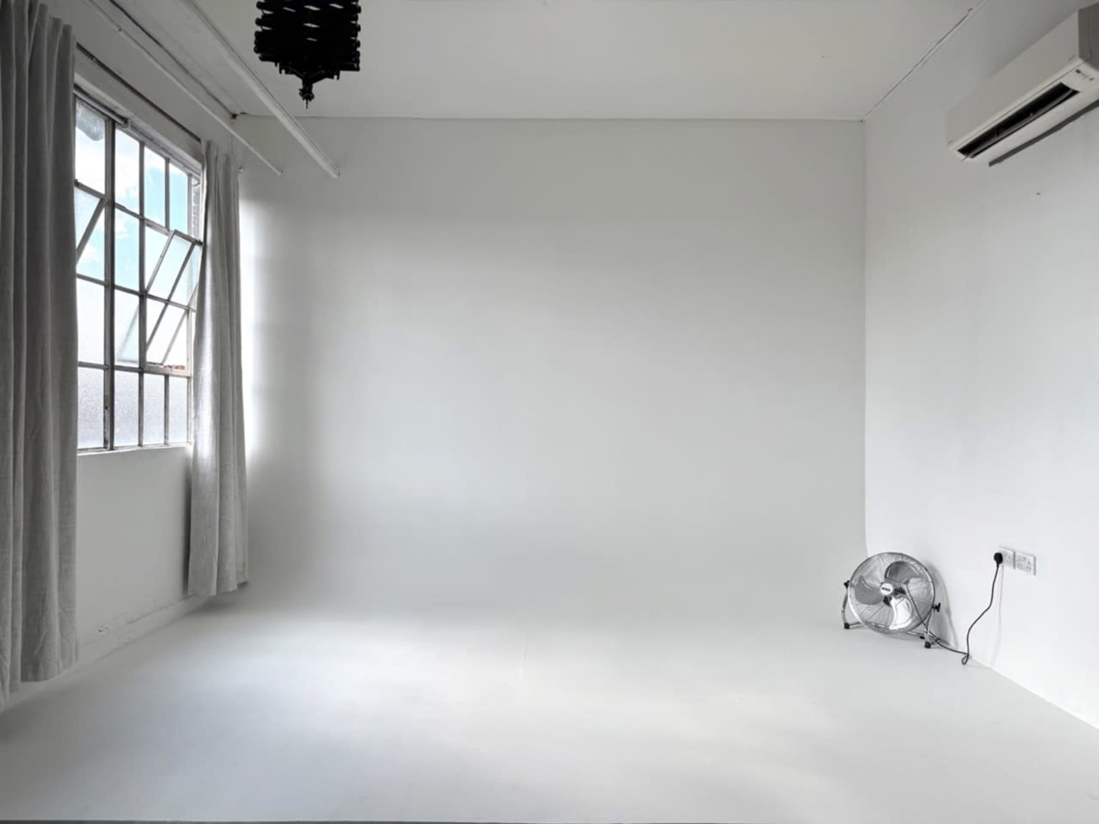 The image shows a minimalist, empty room with a white wall, a window covered by light curtains, and a small fan in the corner.