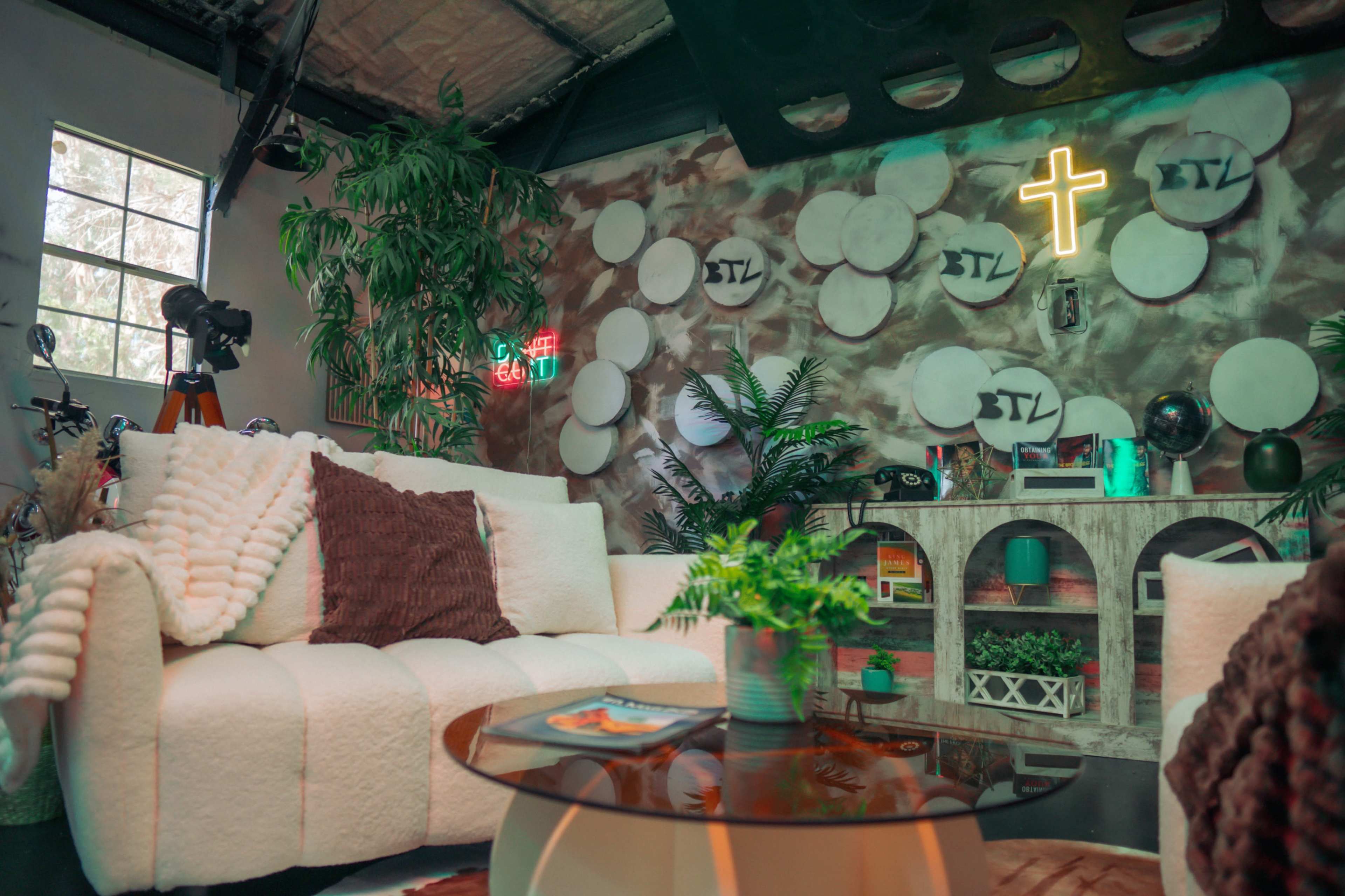 The image shows a contemporary lounge area featuring a white sofa, a glass coffee table, and various decorative elements including plants and a neon cross on the wall.