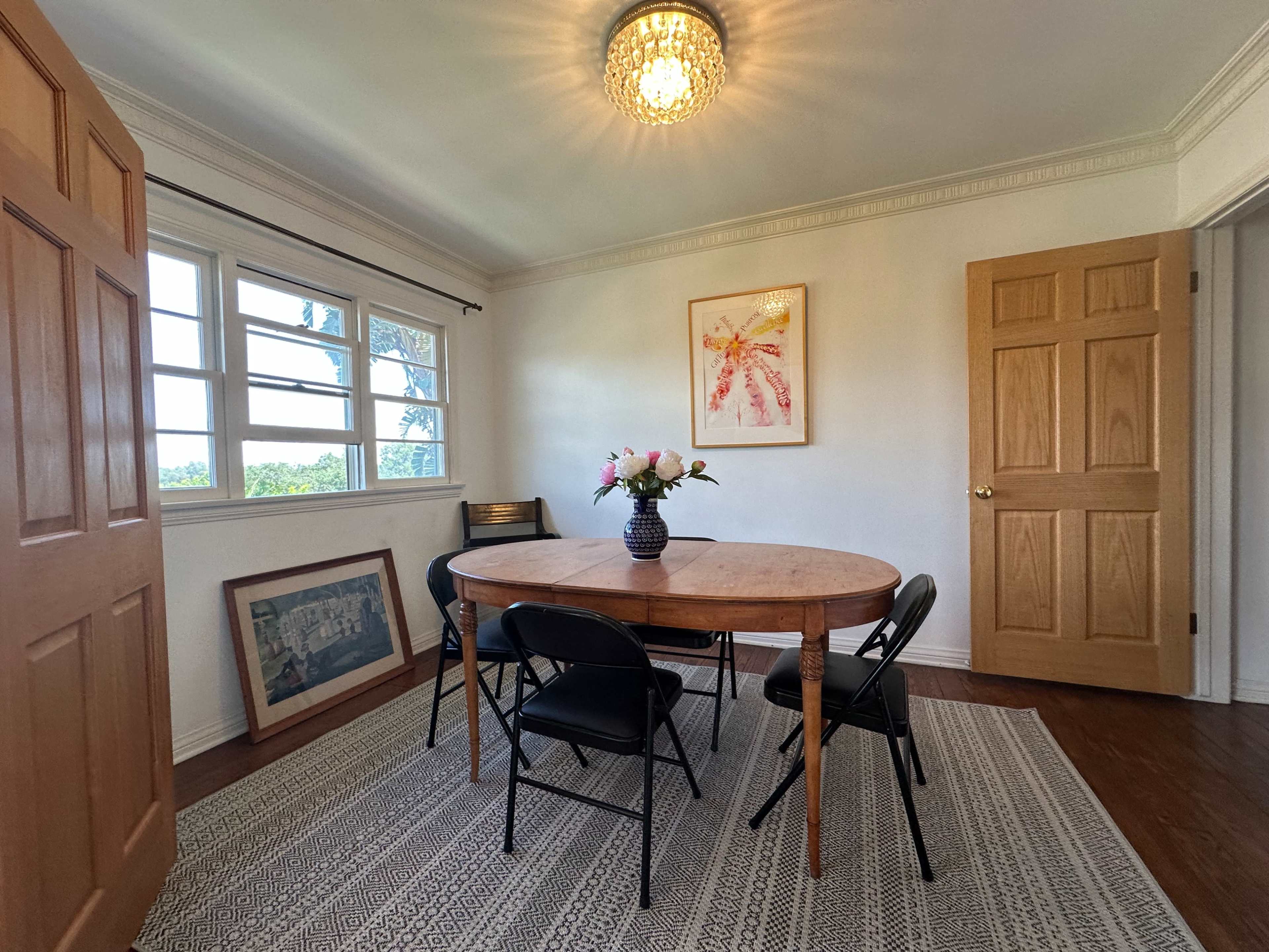 A simple dining room features a round wooden table surrounded by black chairs, with a vase of flowers on the table and artwork hanging on the walls.