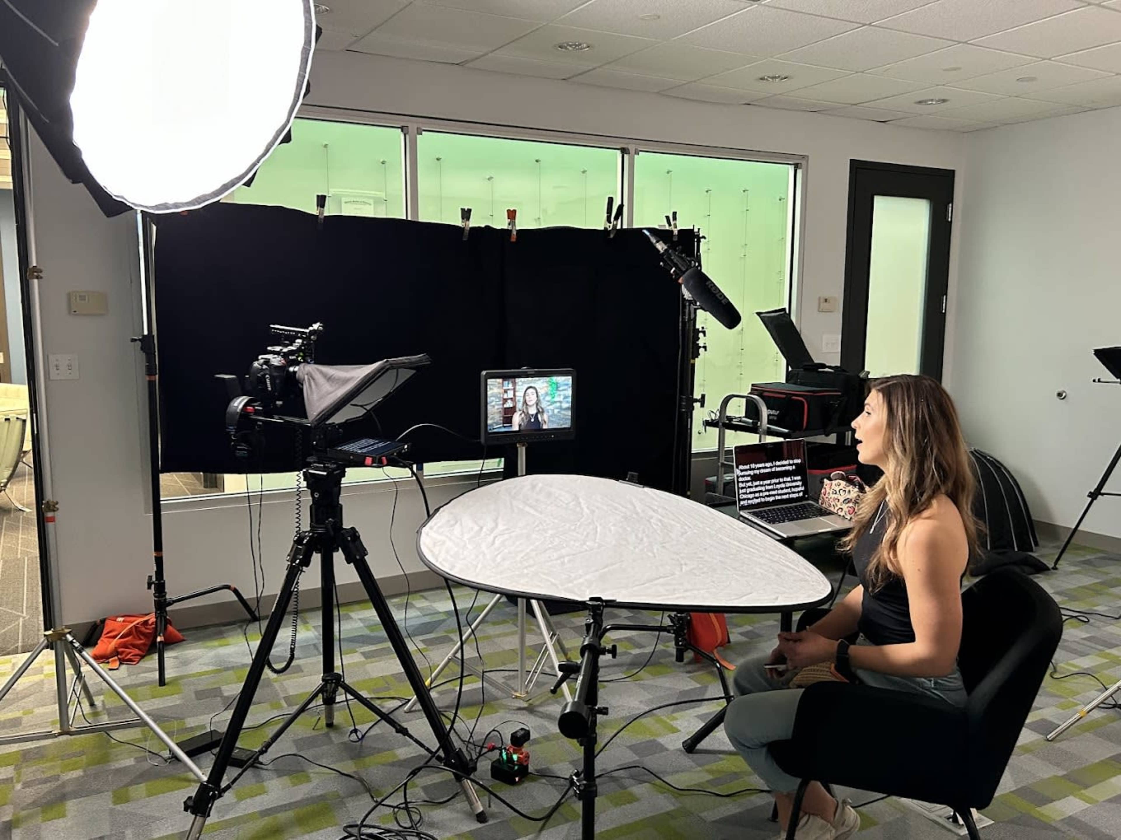 A woman sits in a chair in a recording studio, facing a camera setup with a softbox light and a large backdrop.
