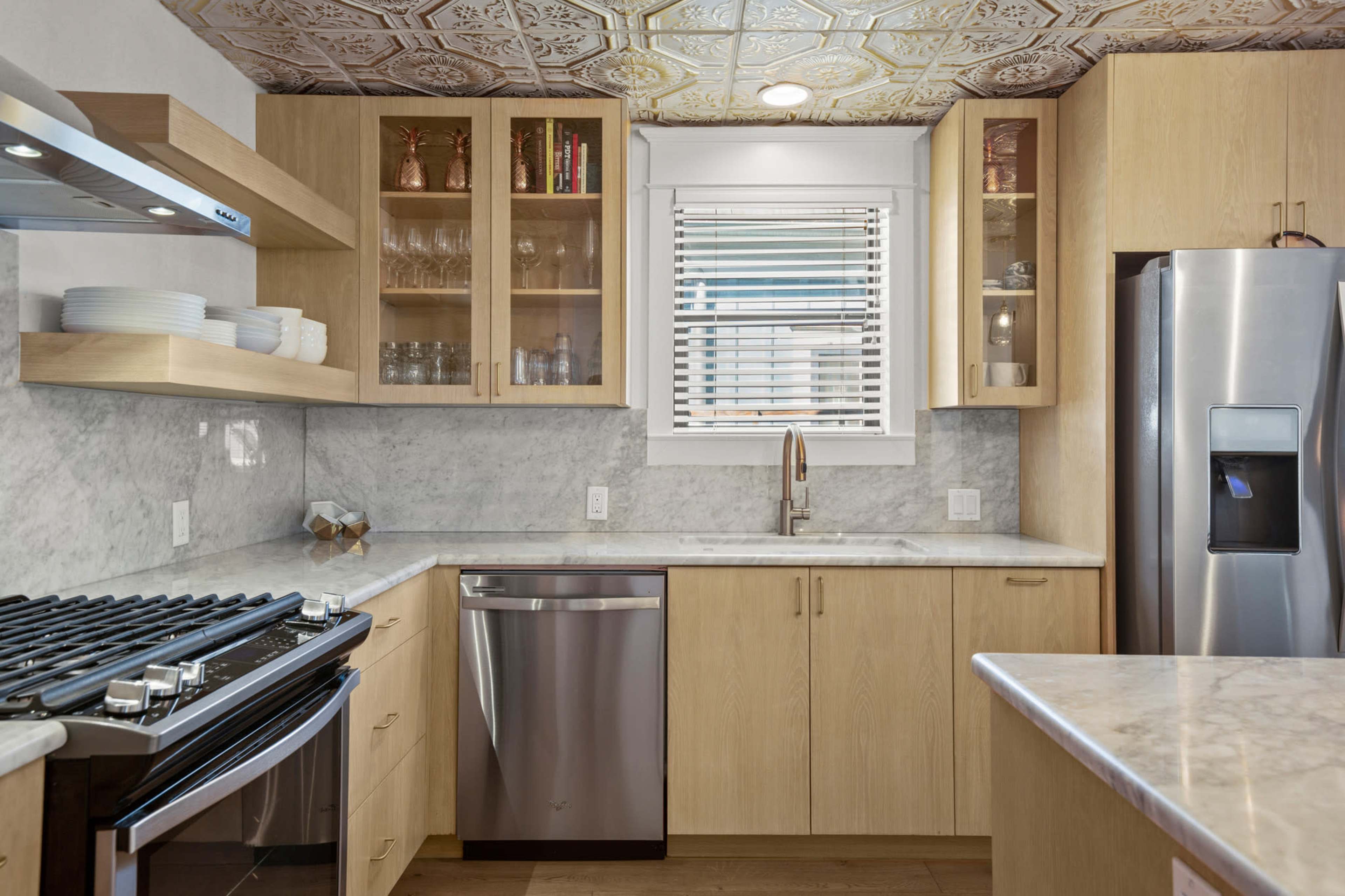 A modern kitchen features light wood cabinets, a marble countertop, stainless steel appliances, and a decorative ceiling.