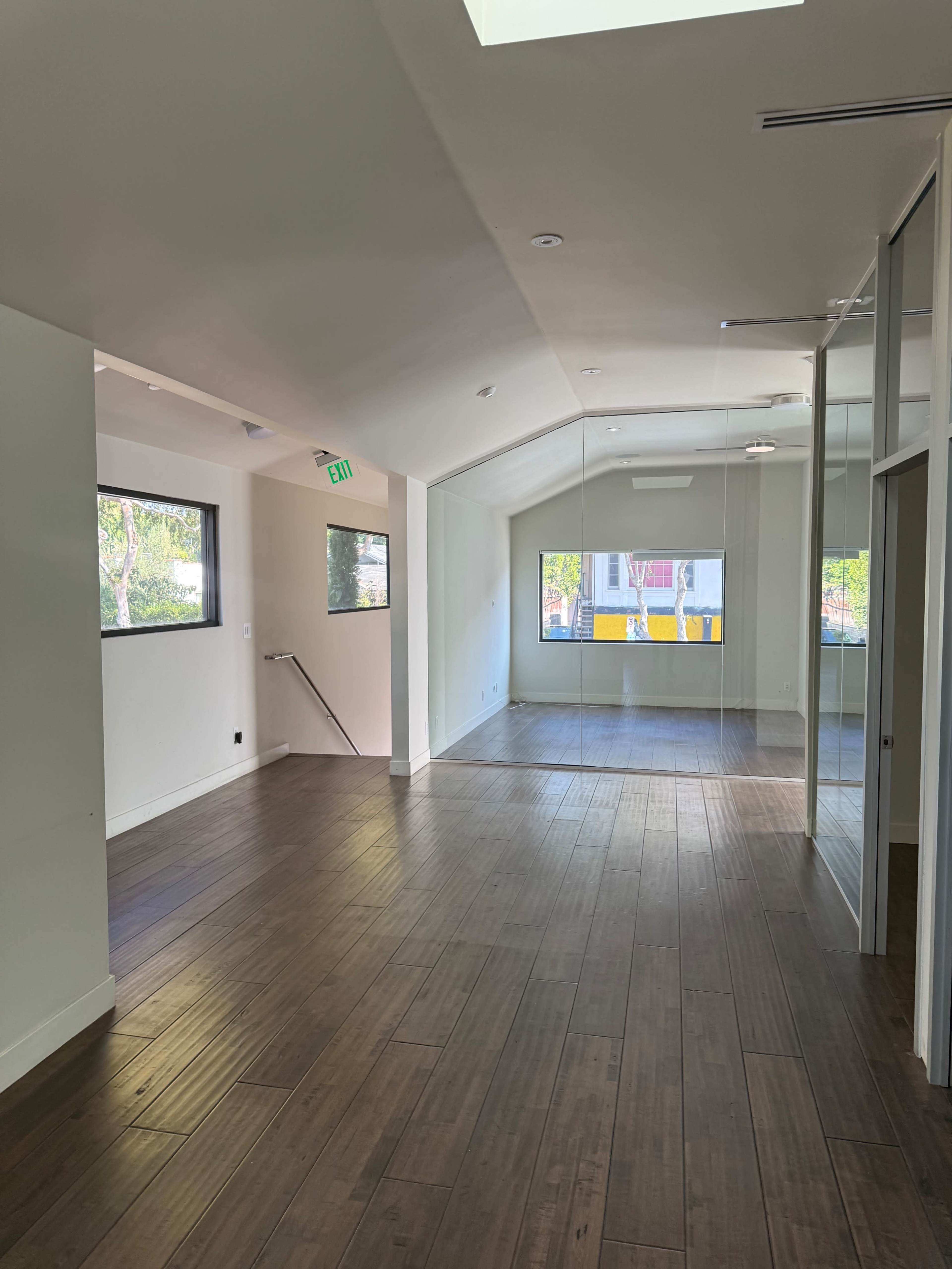 The image shows a spacious, brightly lit interior of an empty room with wooden floors and large windows, featuring a mirrored wall.