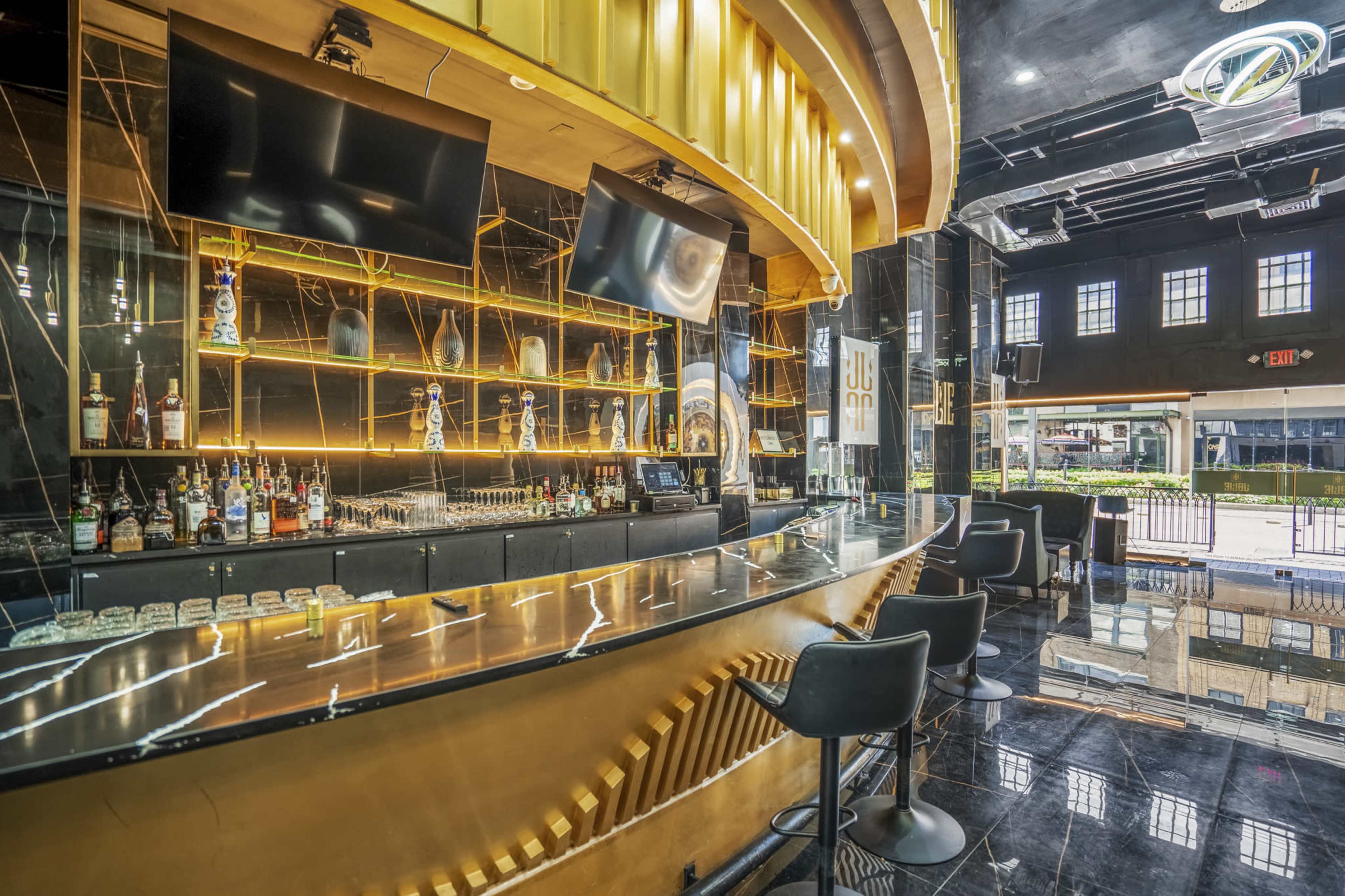 A modern bar area with a sleek curved counter, illuminated shelves featuring a variety of liquor bottles, and large wall-mounted screens.