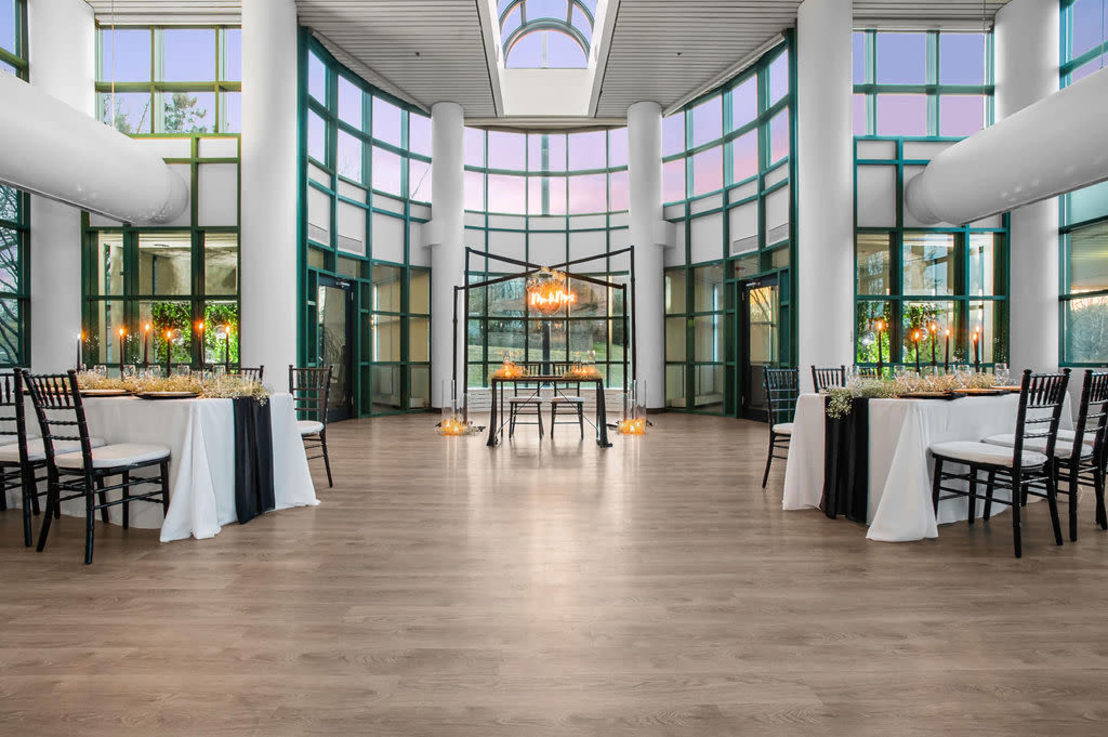 A spacious event room with large windows, two elegantly set dining tables, and a central decorative structure under soft lighting.