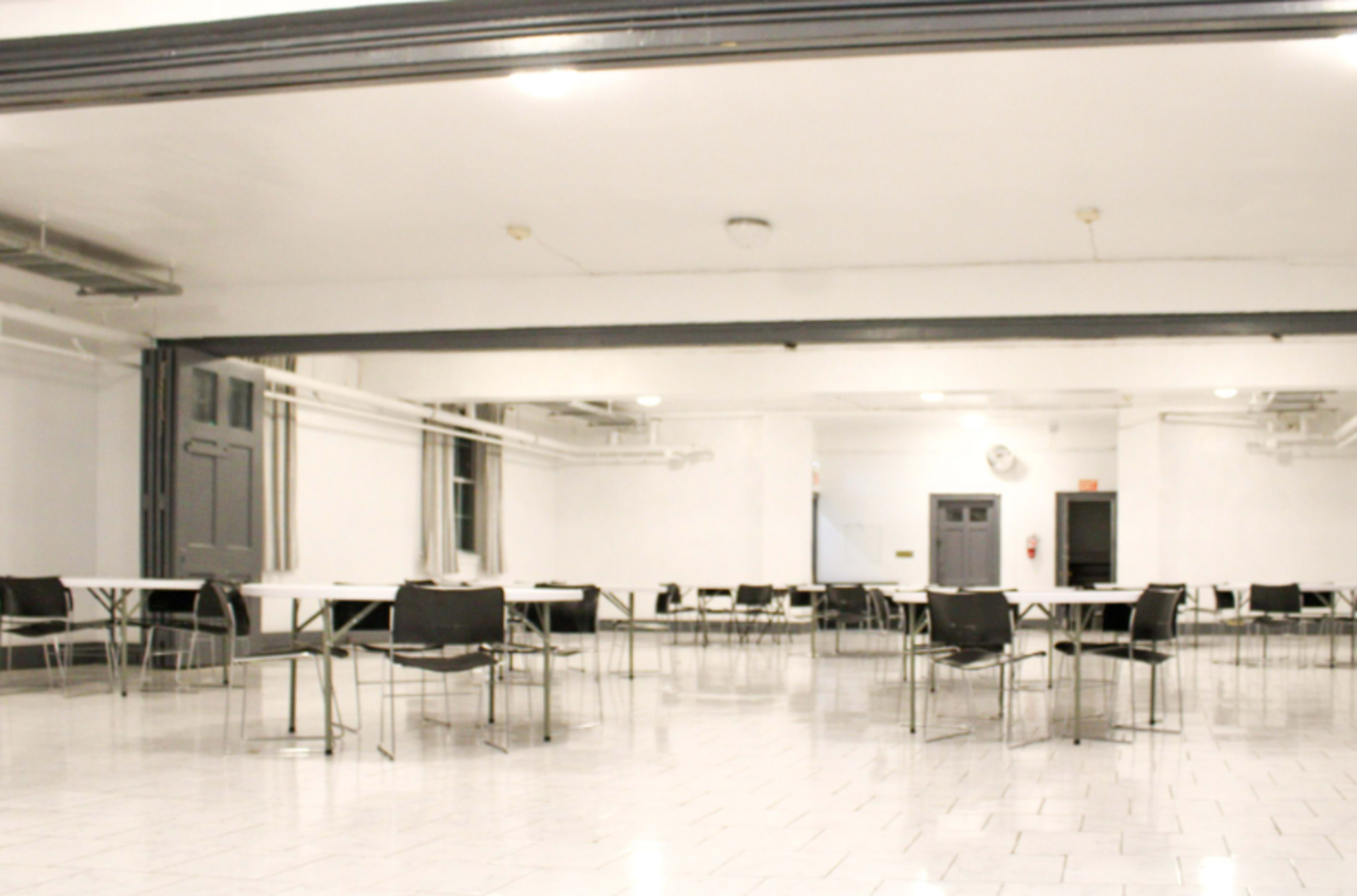 The image shows a spacious, empty room with several tables and chairs arranged in a grid format on a tiled floor.