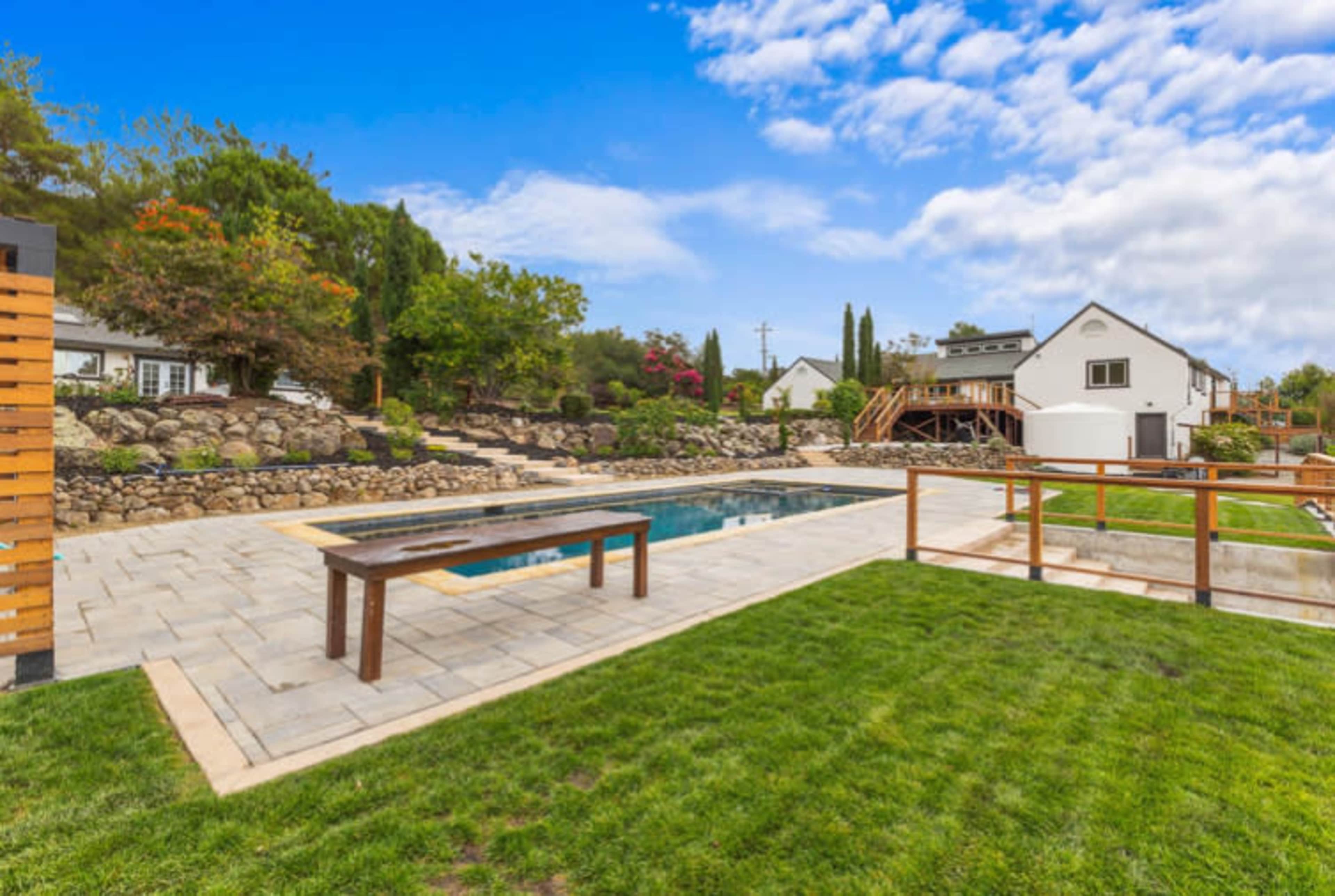 The image depicts a spacious backyard featuring a swimming pool surrounded by a stone patio, a grassy area, and landscaped gardens with trees and shrubs.