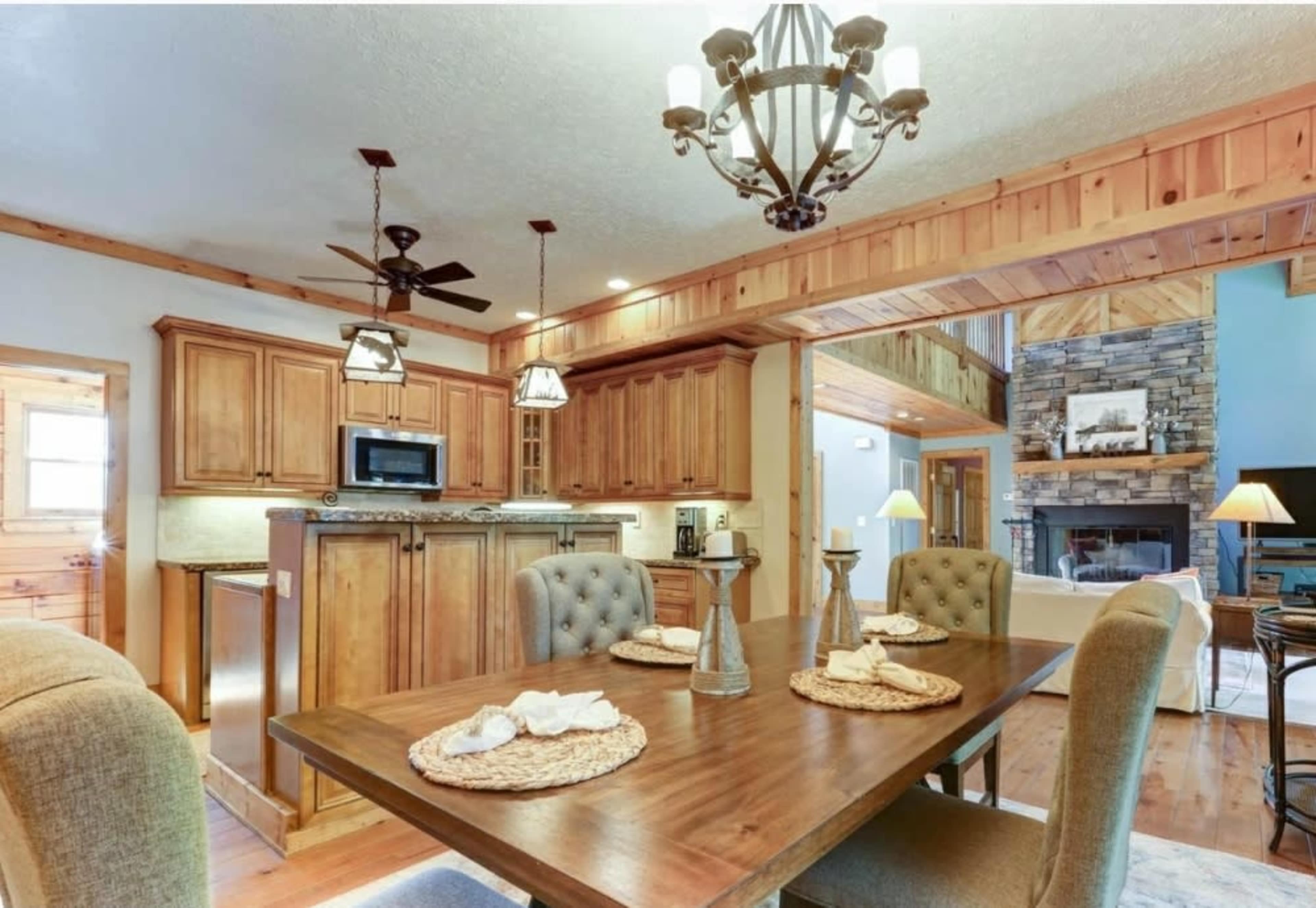 A wooden dining table is set with placemats and utensils, positioned near a kitchen featuring dark wooden cabinets and stainless steel appliances, with a living area visible in the background.
