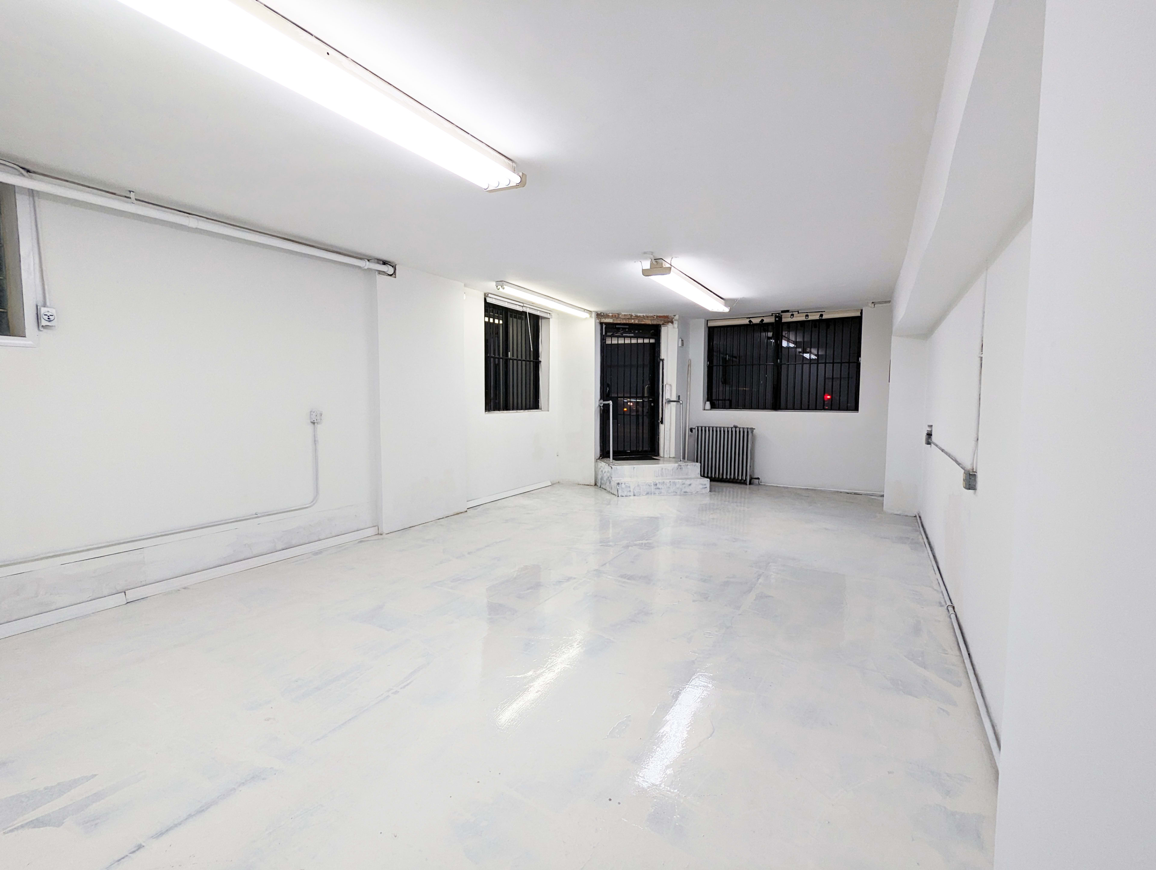 The image shows an empty, white room with a polished floor, featuring windows with metal grates and a staircase at the far end.