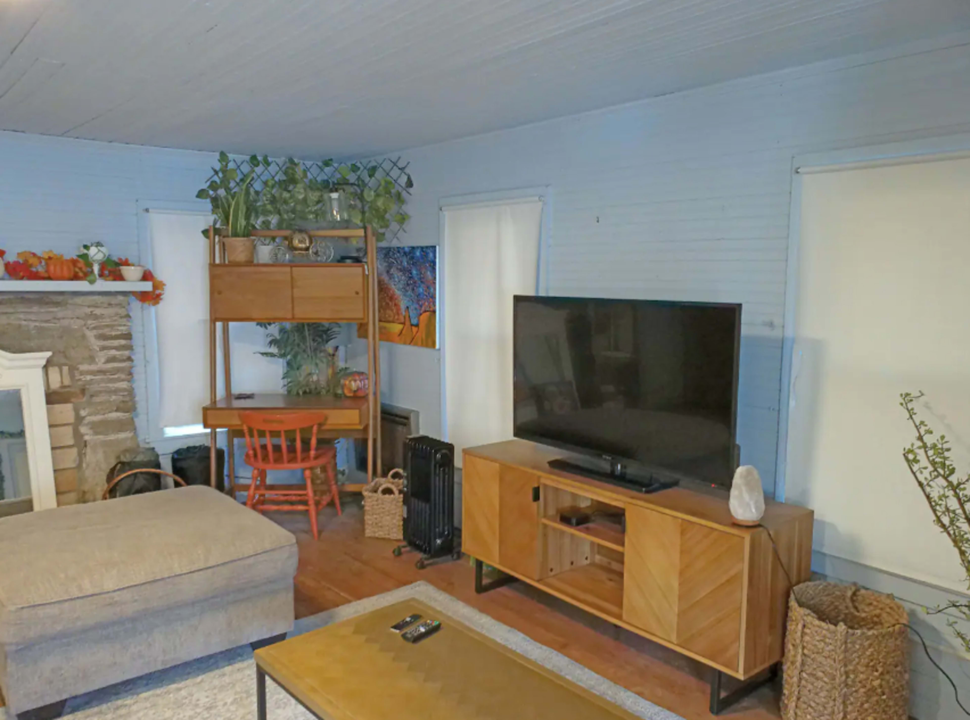 A living room features a sectional sofa, a wooden TV stand with a large television, a desk with a red chair, and decorative plants on shelves against a light blue wall.