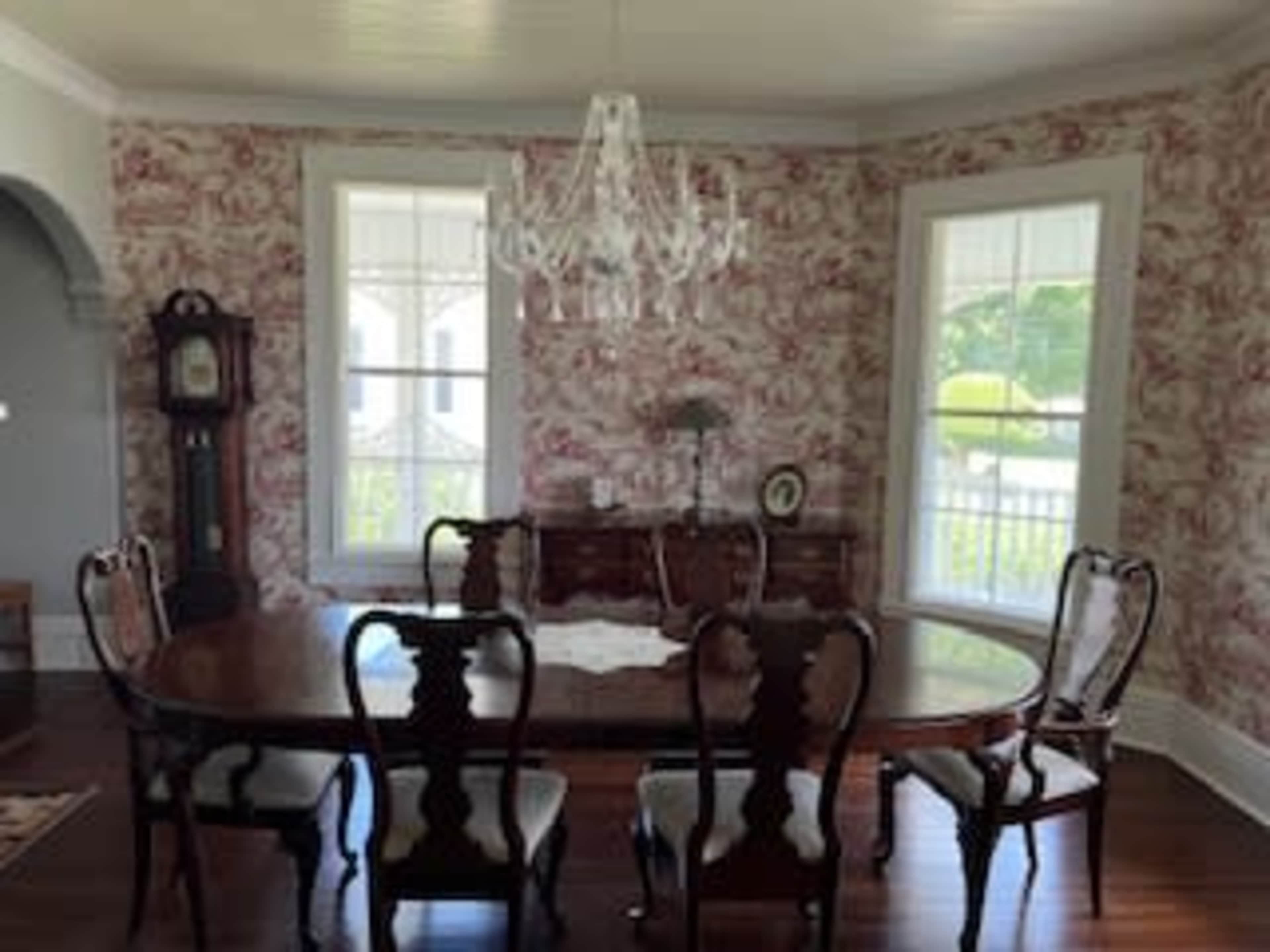 The image features a dining room with a round wooden table surrounded by six chairs, a decorative chandelier overhead, and floral wallpaper.