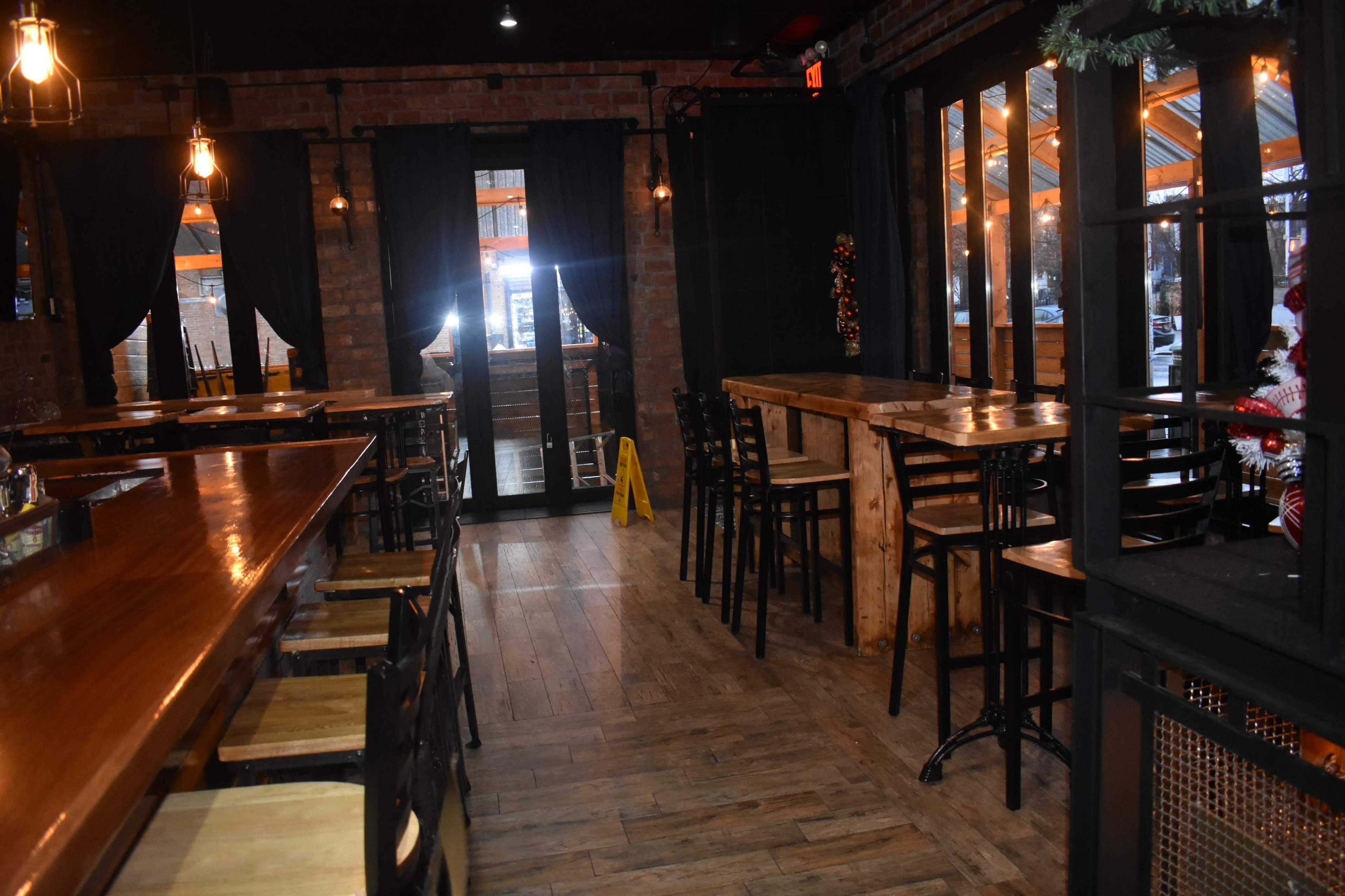 The image shows an empty restaurant with wooden tables and chairs, featuring a bar area and large windows.