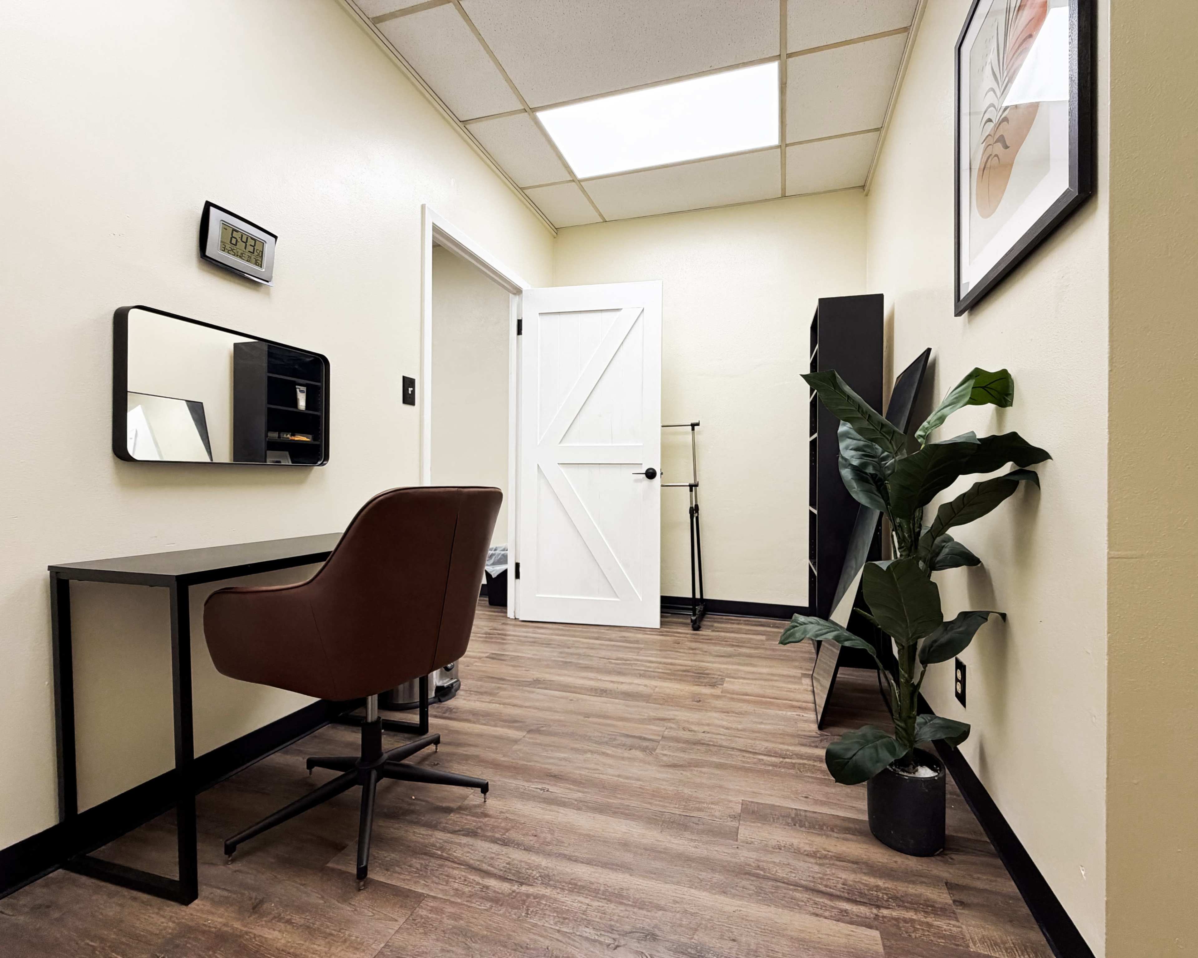 The image shows a small room featuring a desk with a chair, a plant in a pot, and a door leading out, all arranged along the walls.