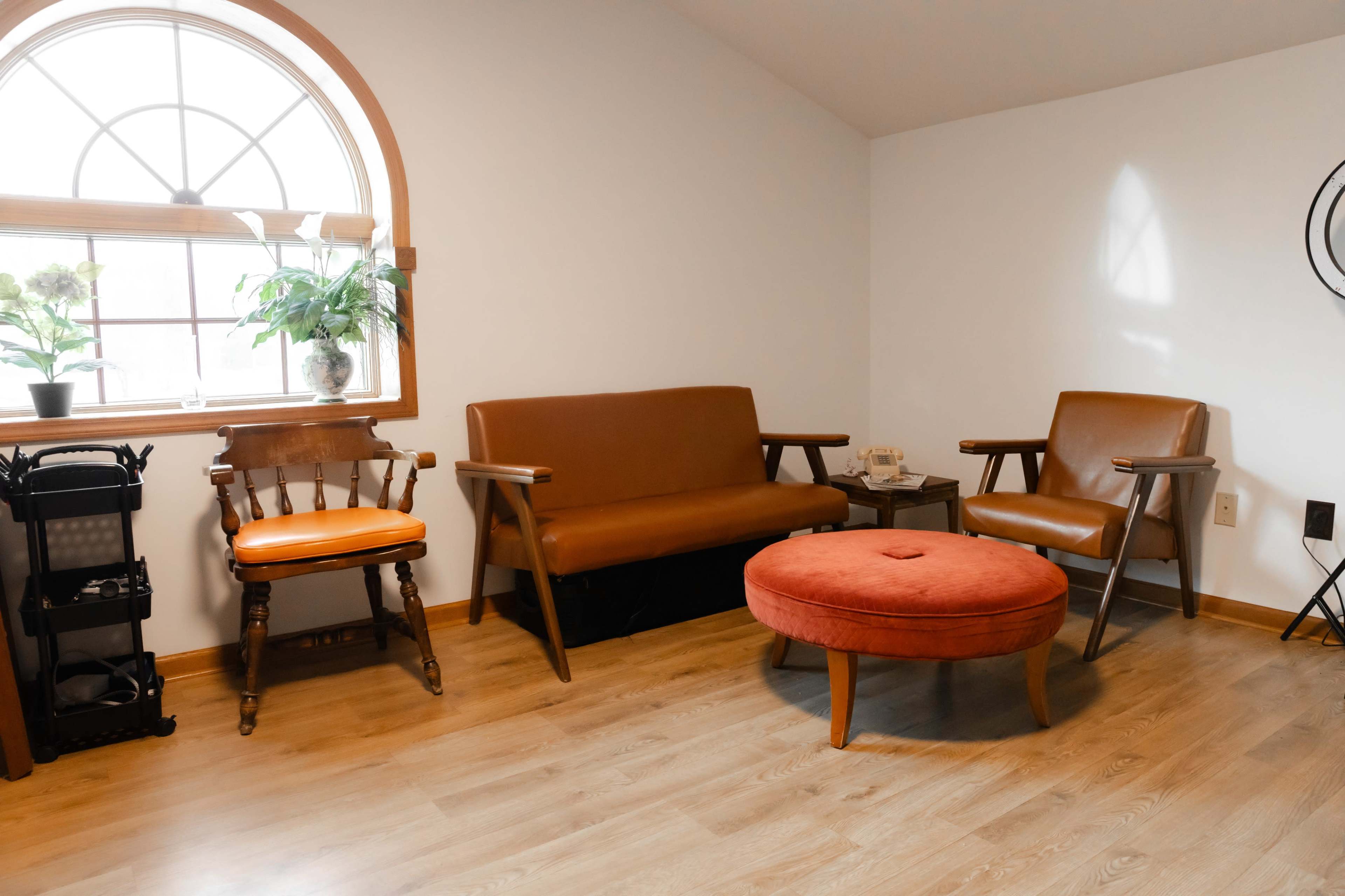 A cozy seating area with a brown sofa, an orange ottoman, and a wooden chair near a window with potted plants.