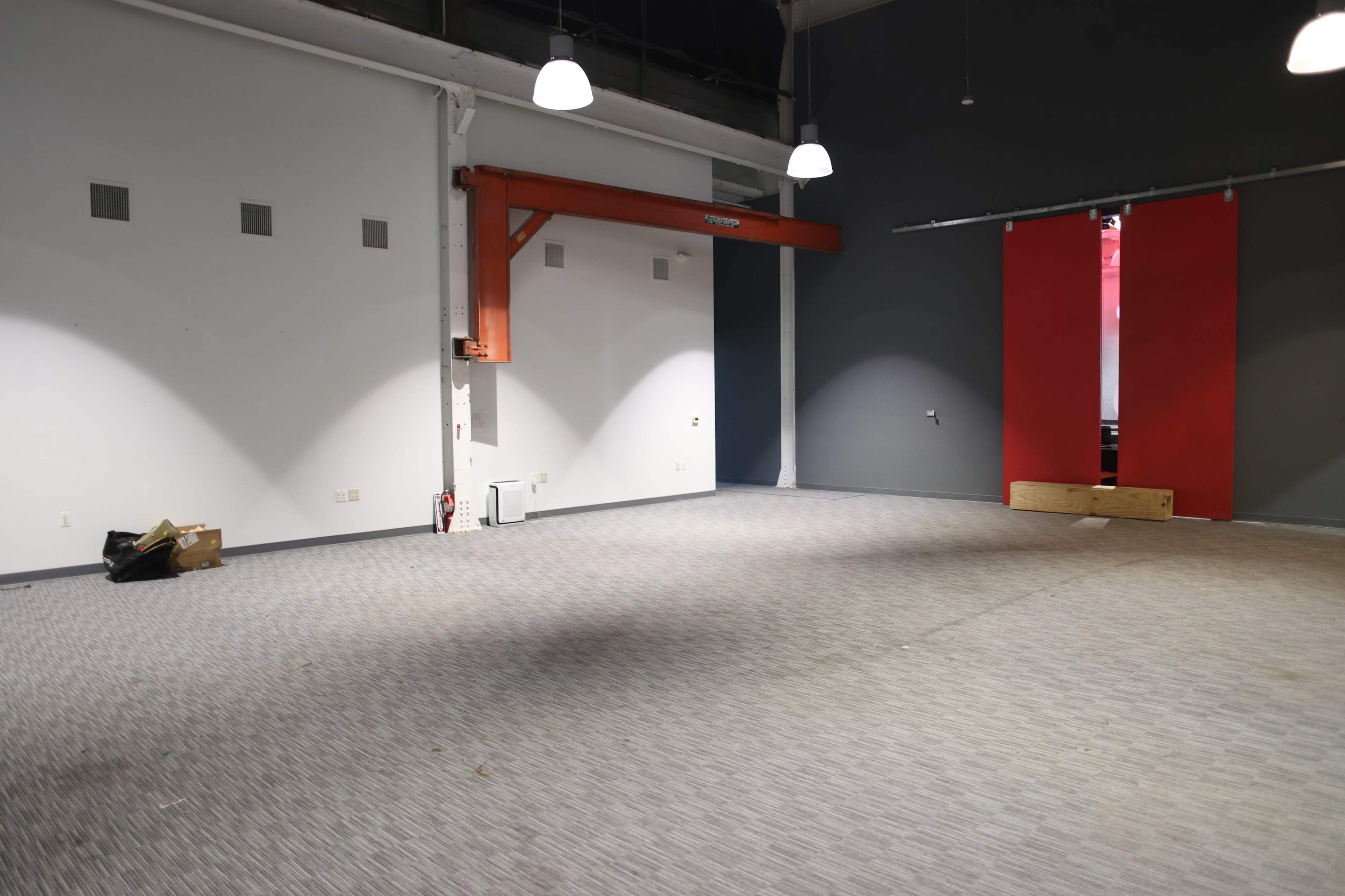 The image shows an empty room with gray walls, a red partition at one end, overhead lights, and a faintly patterned carpet.