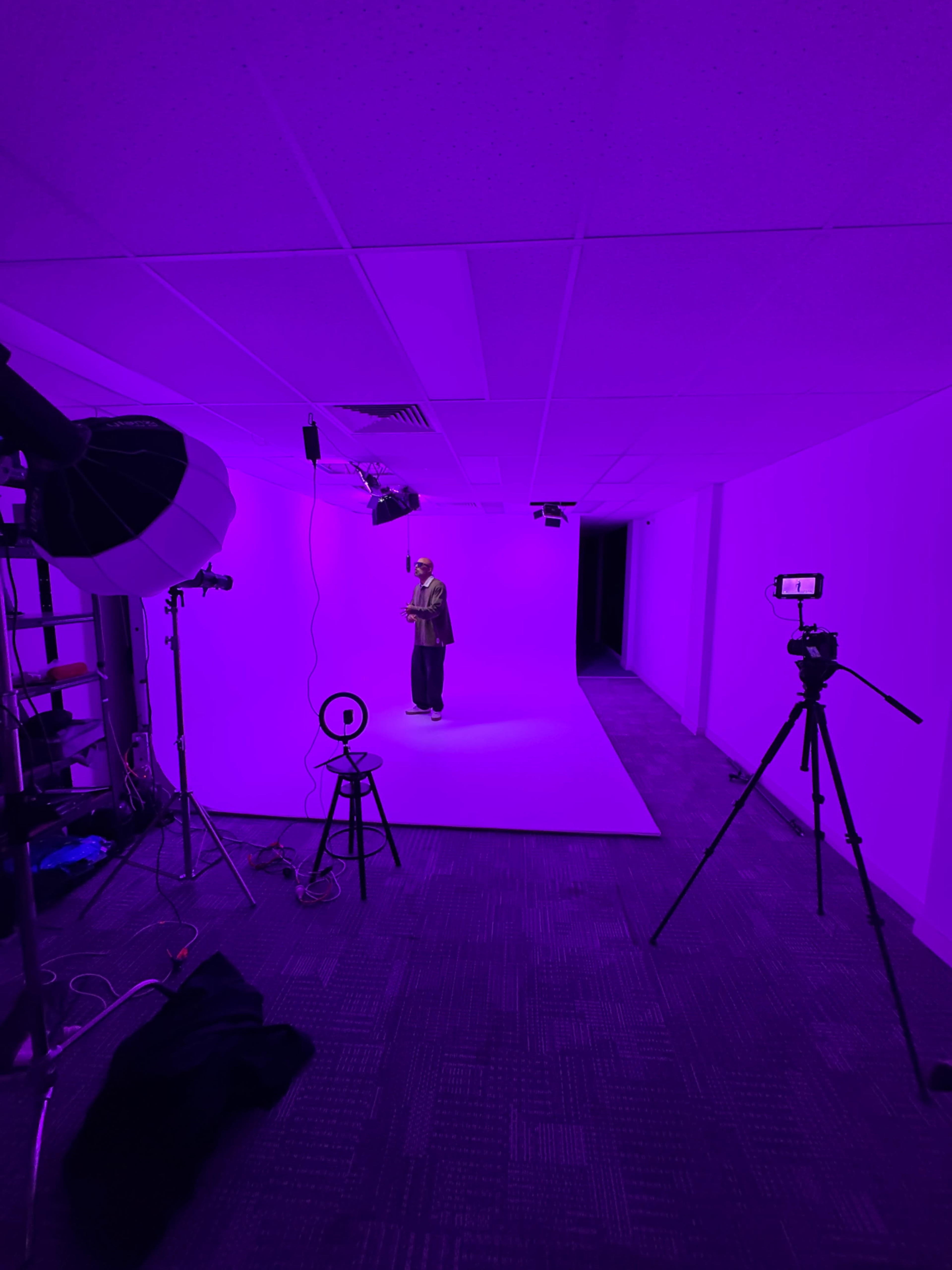 A person is standing on a white backdrop in a brightly lit room with purple walls, surrounded by various photography equipment including a camera on a tripod and a softbox light.