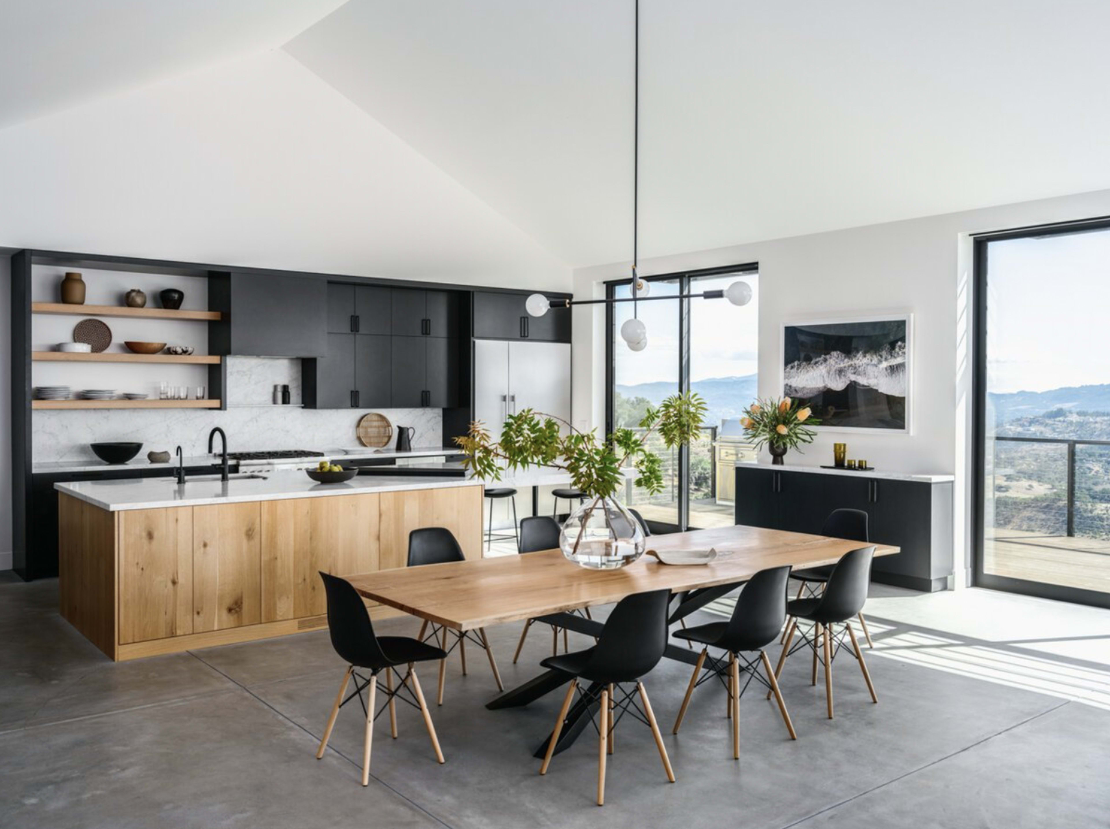 A modern kitchen features a central wooden dining table surrounded by black chairs, with sleek black and white cabinetry and large windows showcasing a scenic view.
