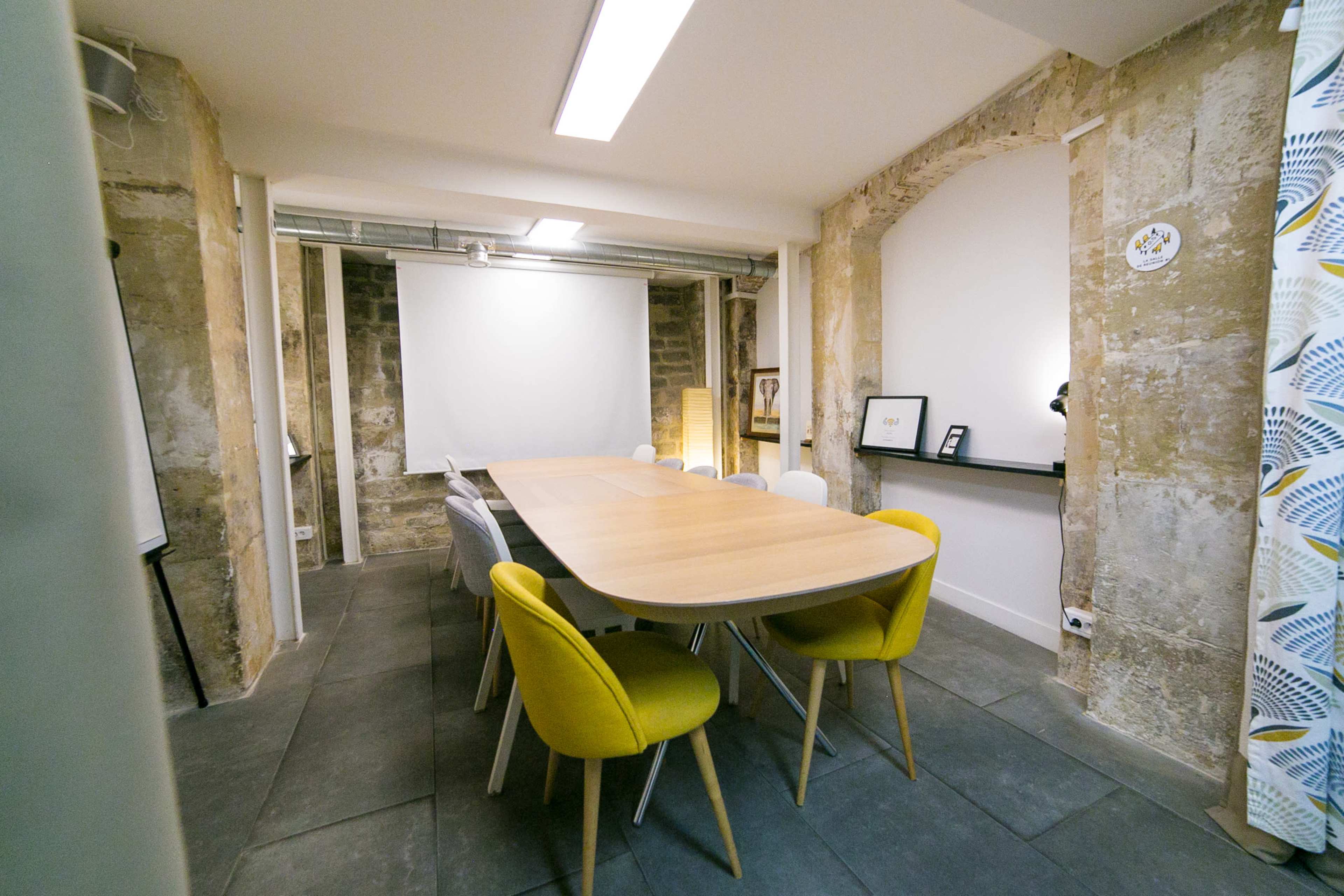 A modern conference room features a long wooden table surrounded by white and yellow chairs, with a projector screen at one end and exposed stone walls.