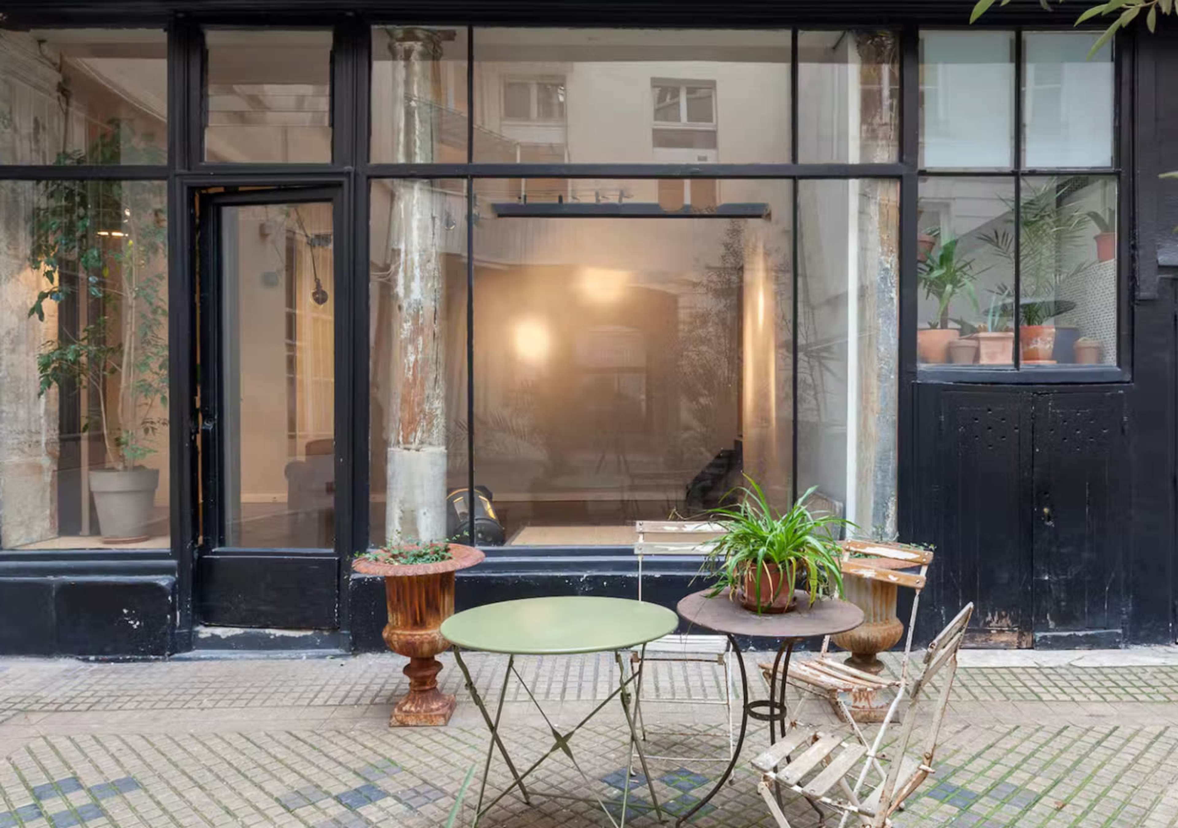 A small outdoor seating area with a green table and two metal chairs is set in front of a large glass storefront featuring plants and minimal interior decor.