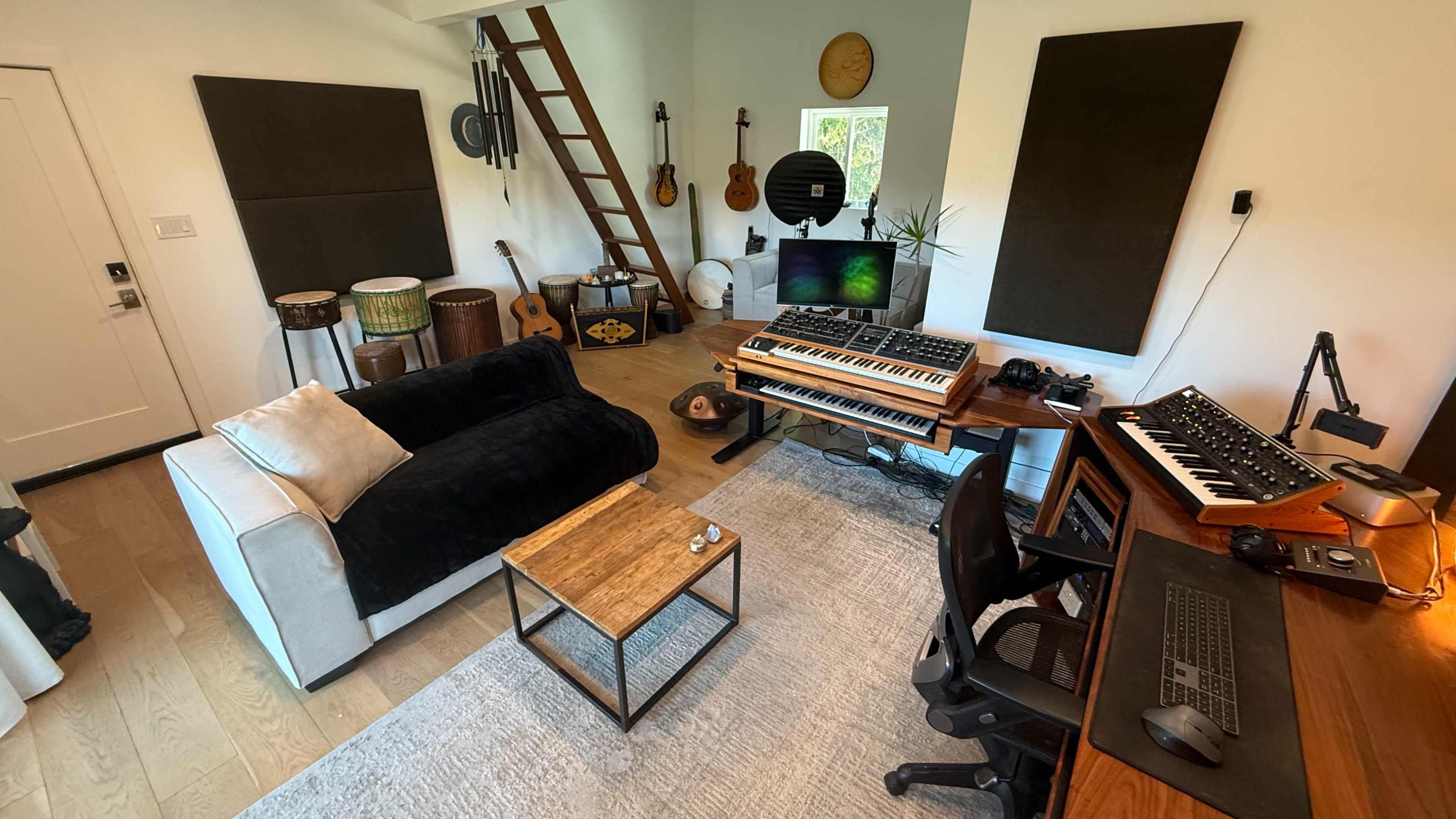 The image depicts a music studio featuring a black sofa, a wooden desk with music equipment, and various musical instruments arranged throughout the space.