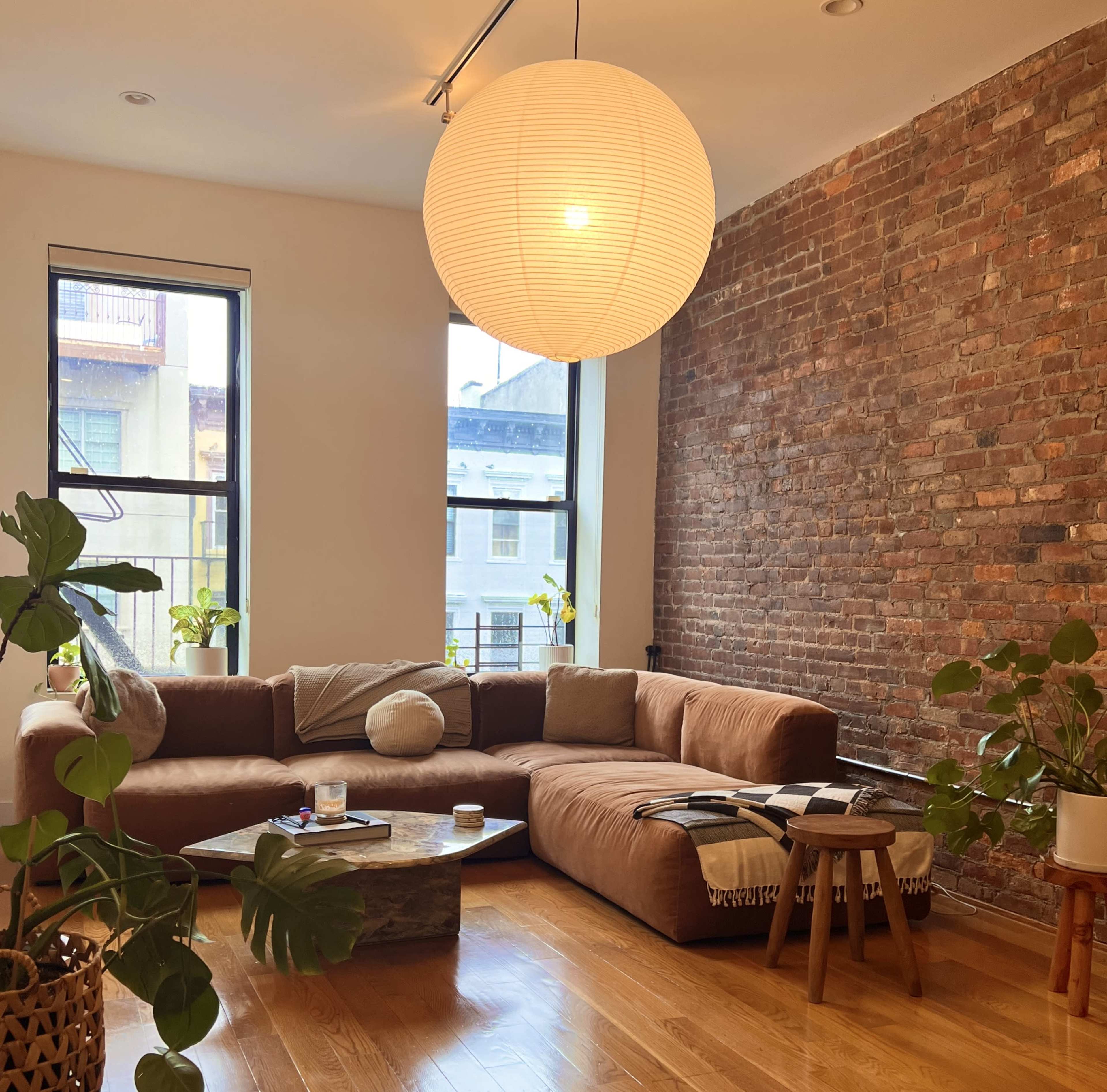 A living room with a brown sectional sofa, a round coffee table, a large paper lantern light fixture, and several plants against a brick wall.