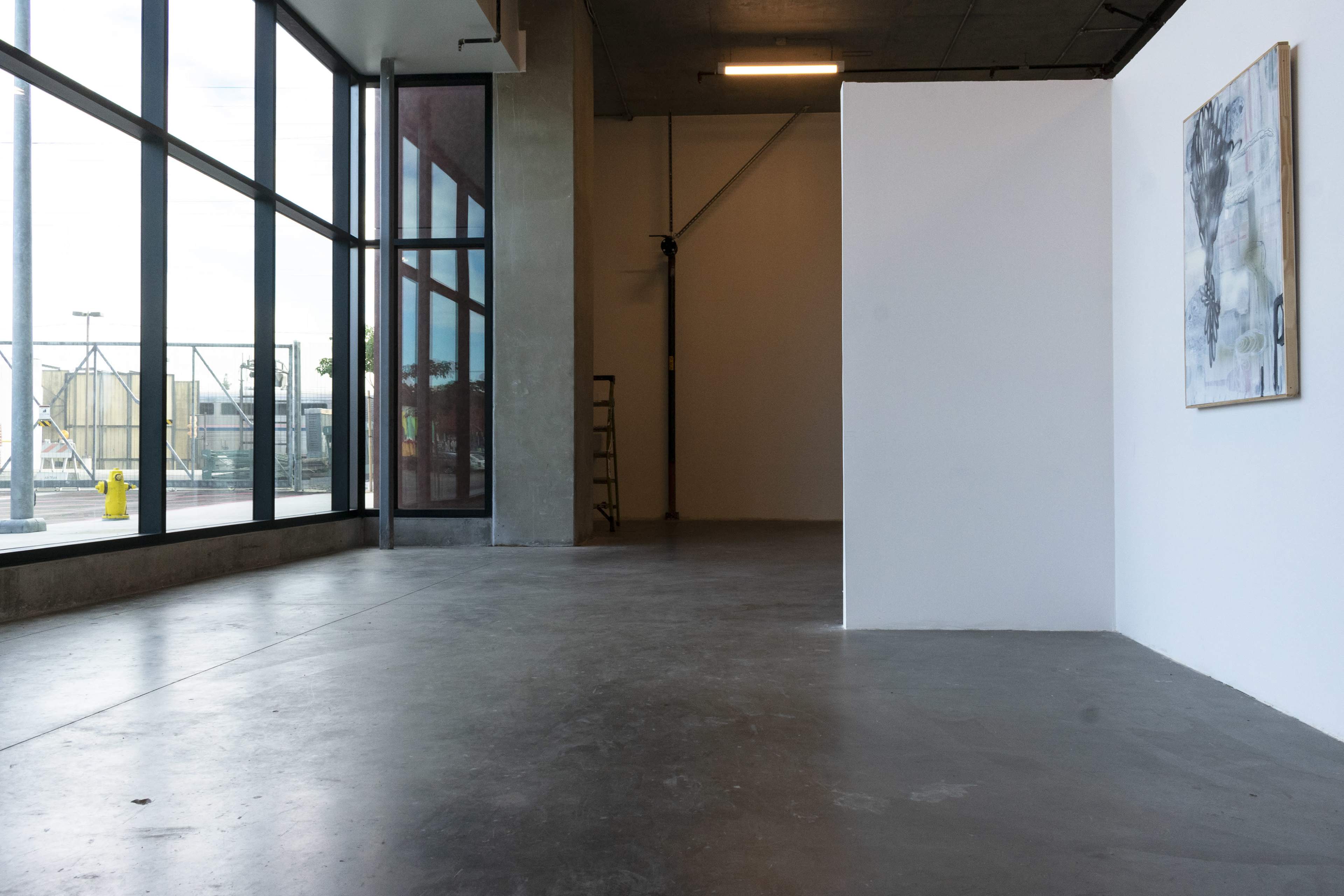 A spacious, minimalist art gallery with concrete floors, large windows, and a single artwork on the wall.