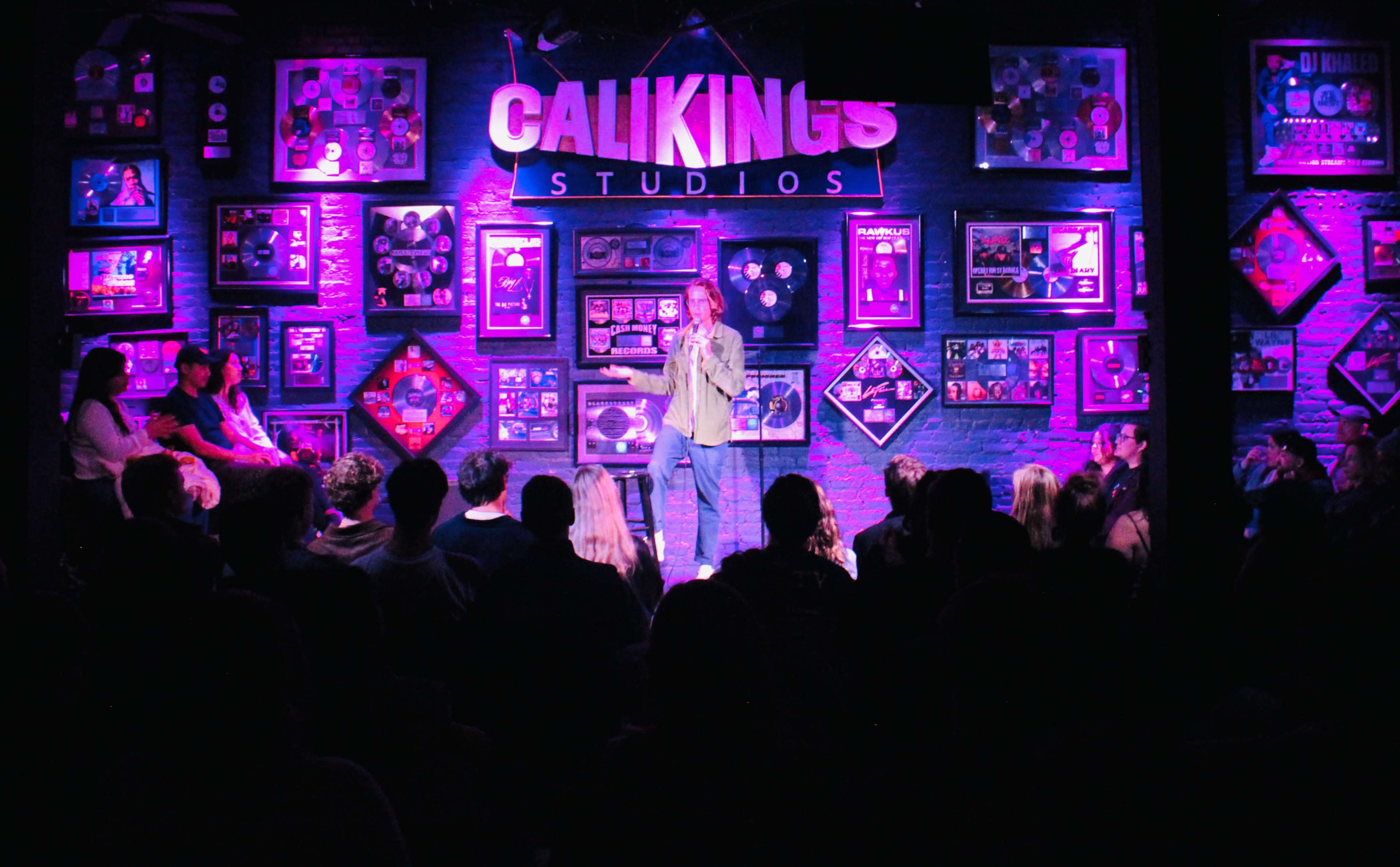 A comedian performs on stage at Calikings Studios, surrounded by framed records and awards, while an audience watches.