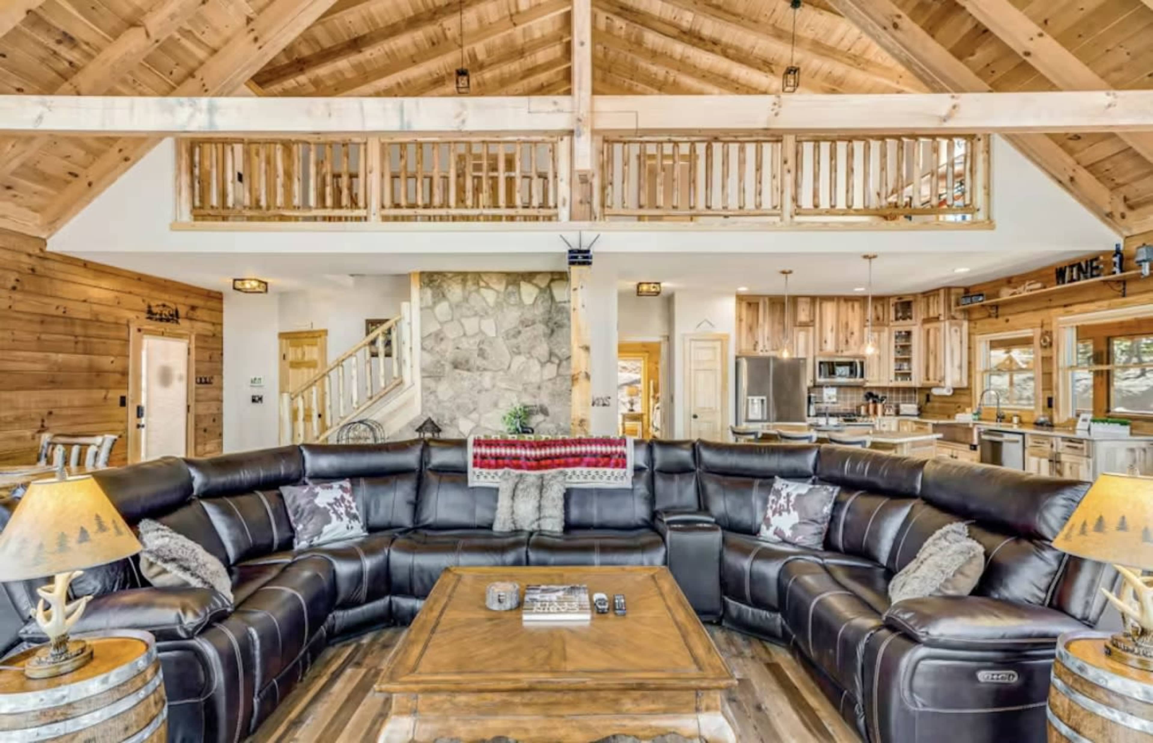 The image shows a spacious, wooden interior of a cabin living room featuring a large sectional sofa, a stone fireplace, and an open staircase leading to a loft area.