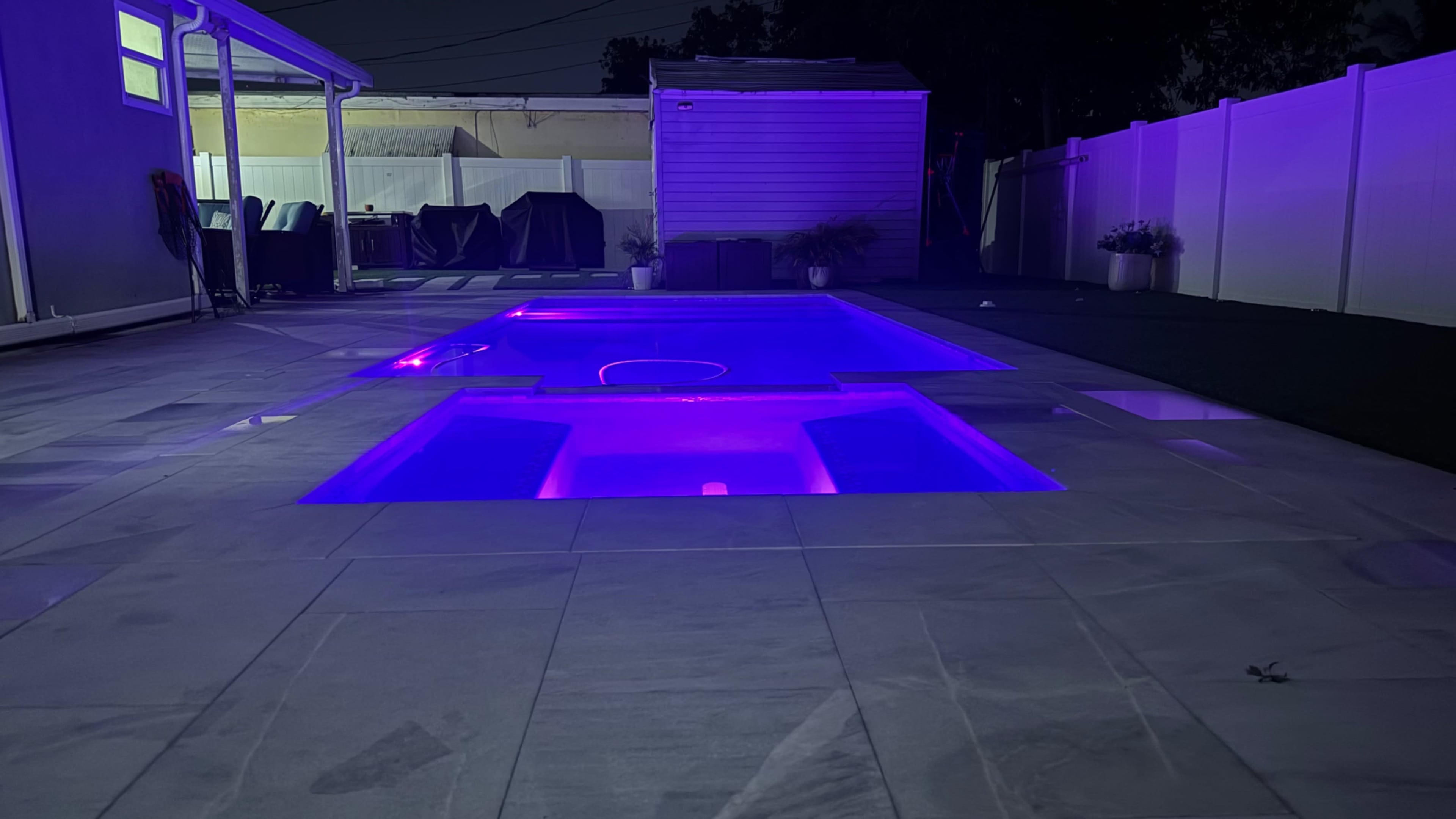 The image shows a backyard at night with a pool illuminated by purple lights, surrounded by a stone patio and a fenced area.