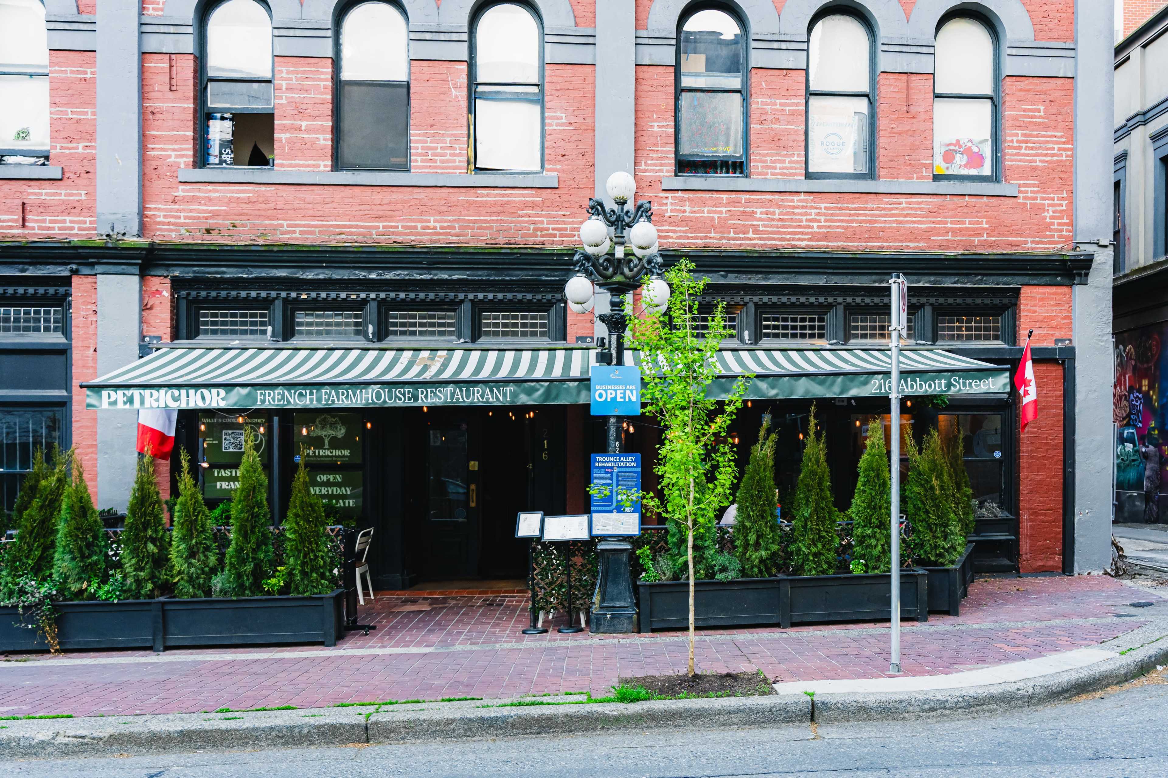 The image shows the exterior of a French farmhouse restaurant called "Petrichor," featuring a striped awning, potted greenery, and a sign indicating it is open, located on a street with brick buildings.