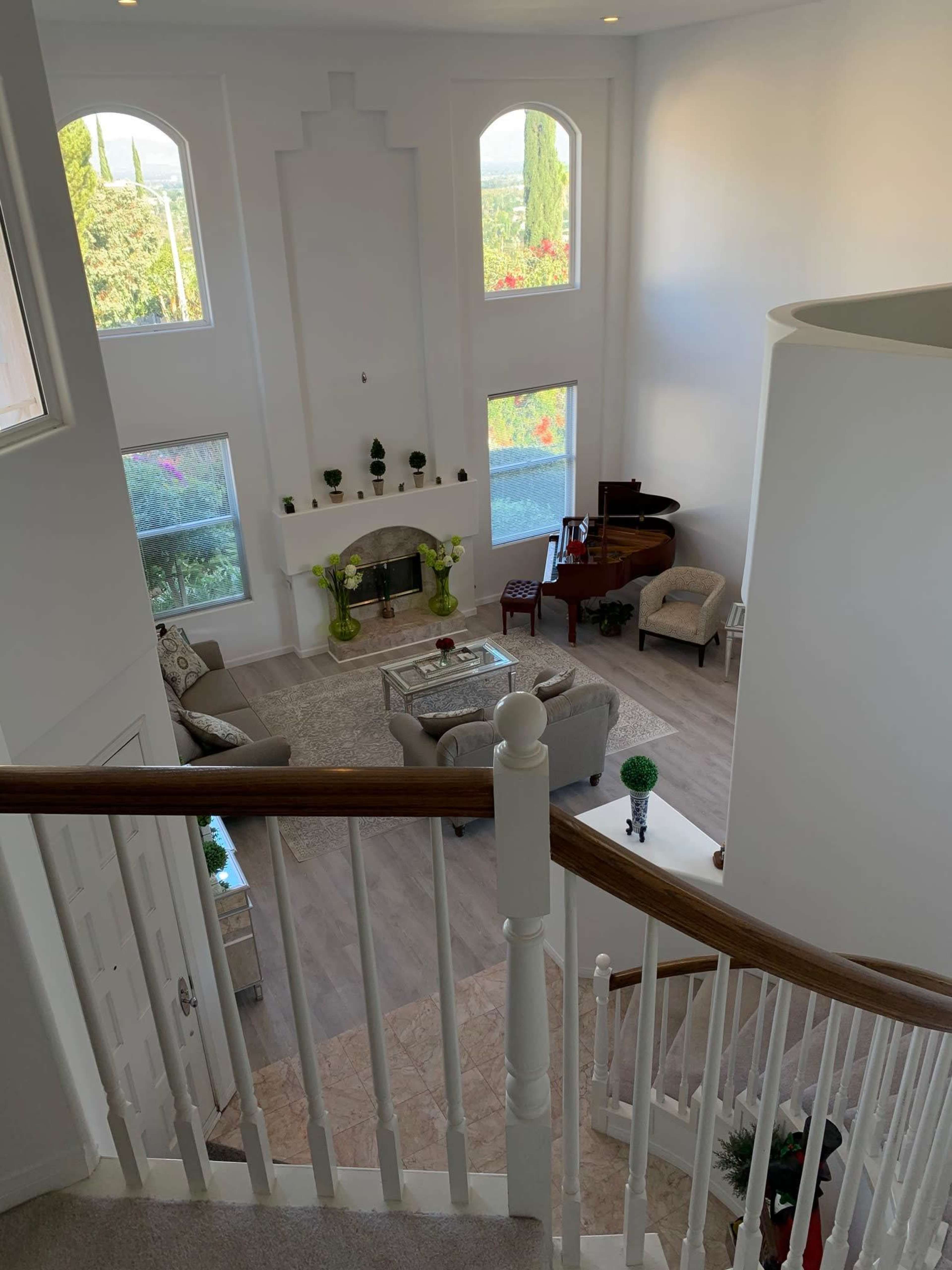 A spacious living room with a fireplace, piano, and large windows overlooks a garden view, seen from an upper landing.