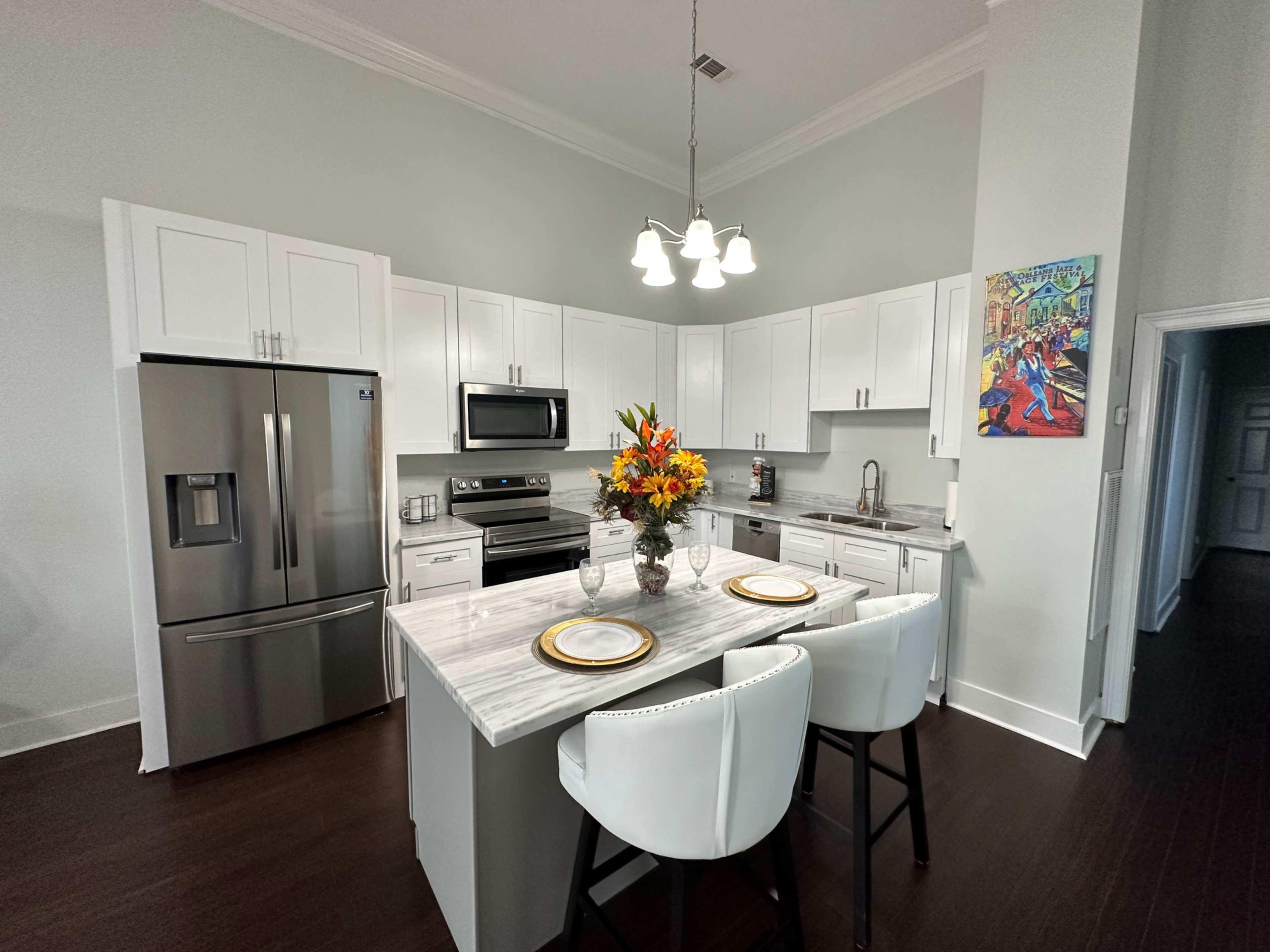 A modern kitchen with white cabinetry, stainless steel appliances, a marble countertop island with two bar stools, and a vase of flowers on the island.