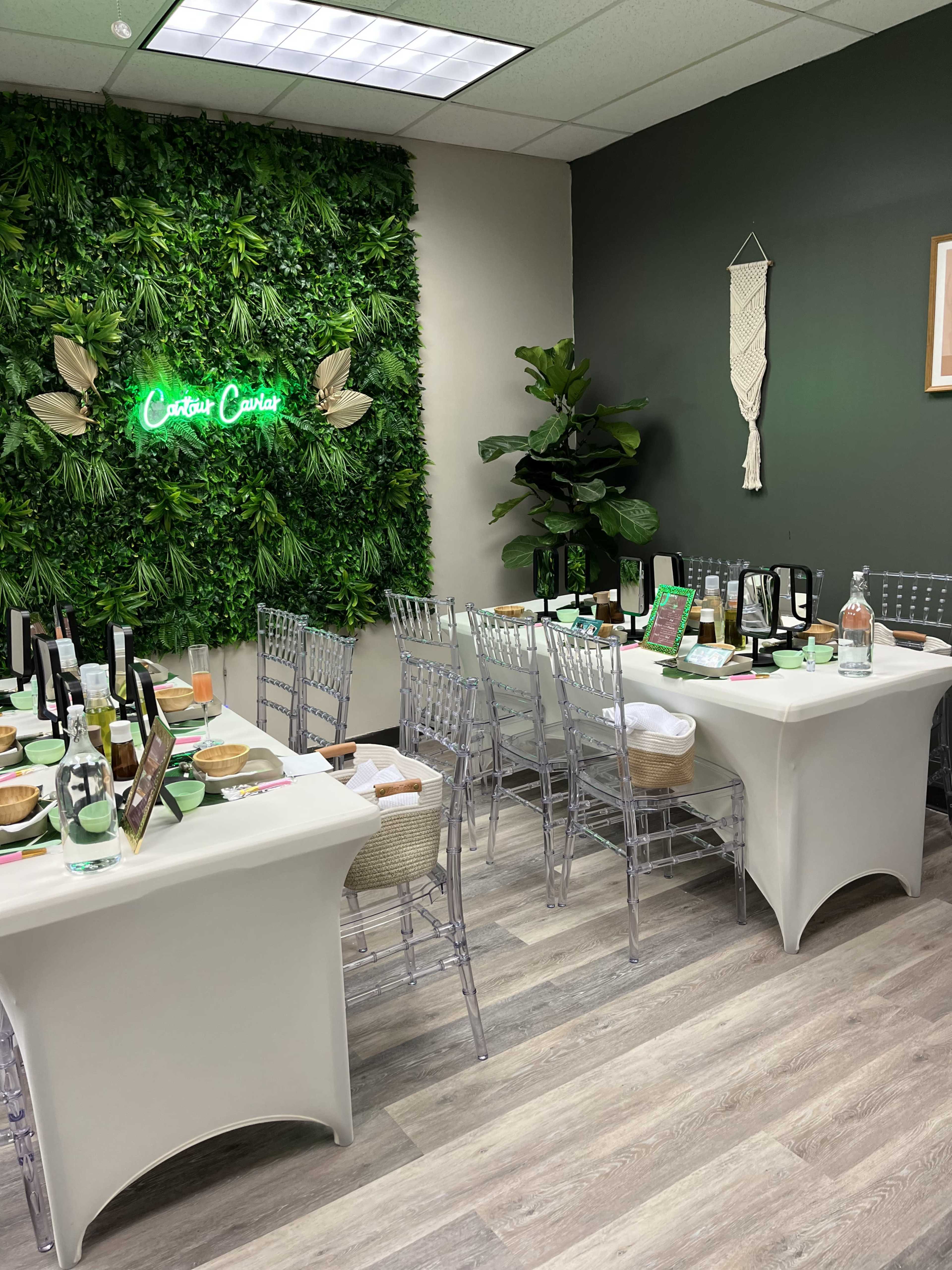 A dining setup with clear chairs, a green foliage wall, and a neon sign, arranged for a gathering.