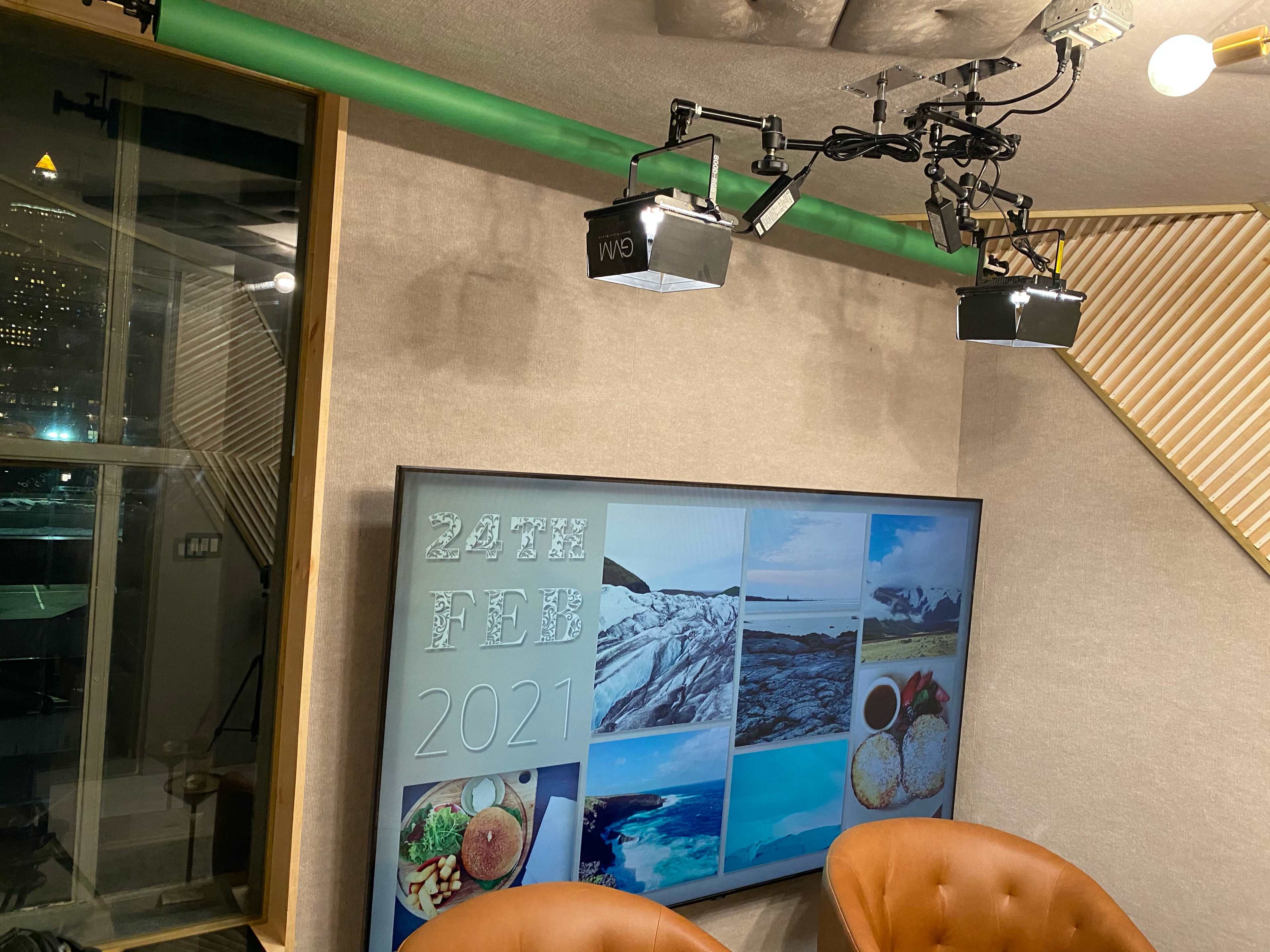 A well-lit studio setup featuring a large screen displaying a collage of photographs and a date, "24th FEB 2021," with two brown chairs positioned in front.