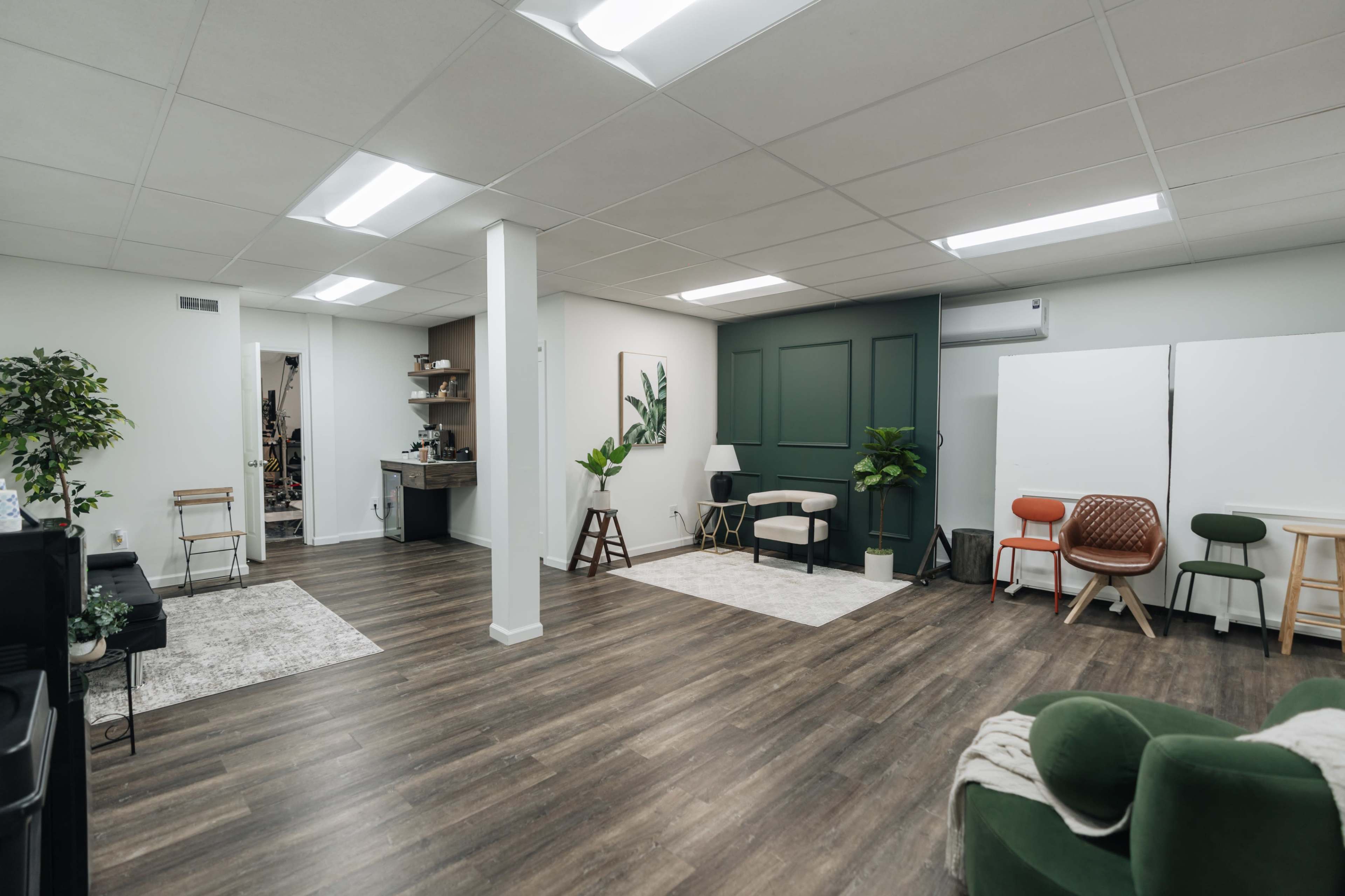 The image shows a spacious, well-lit room with various seating arrangements, wooden flooring, and plants, featuring a green accent wall.